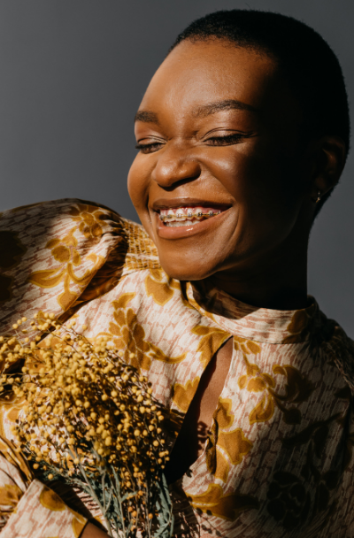 A woman smiling and holding a bouquet of yellow flowers, wearing a floral top with puffed sleeves, against a gray background.