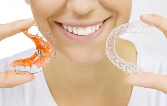 Person holding a clear orthodontic retainer in one hand and a colorful braces bracket in the other, smiling.