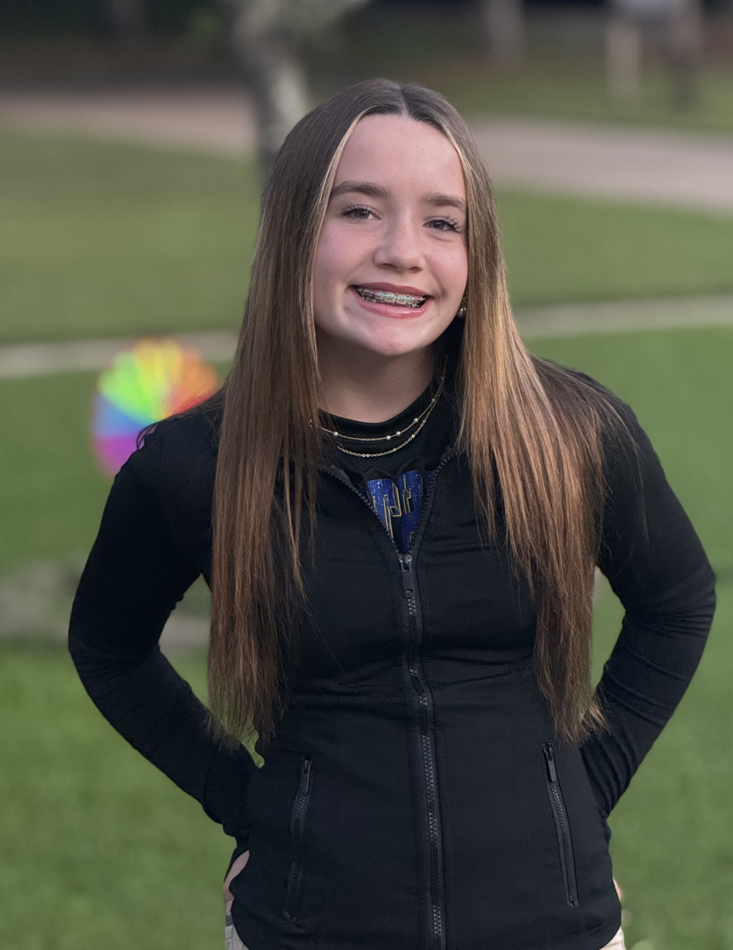A smiling girl with long straight hair, braces, wearing a black zip-up jacket and layered necklaces, standing outdoors on grass with a blurred colorful ball in the background.