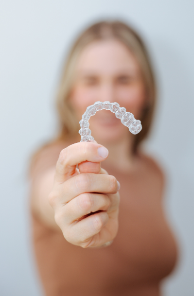 A woman with blonde hair holding a clear orthodontic retainer in front of her face.