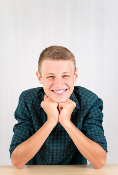 A young boy with short hair smiling and resting his chin on his hands, wearing a green and blue checked shirt.