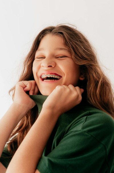 A girl with long brown hair and braces, smiling and holding her green shirt collar.