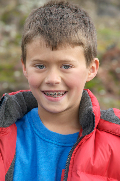 Young boy smiling outdoors, wearing a red jacket and braces.