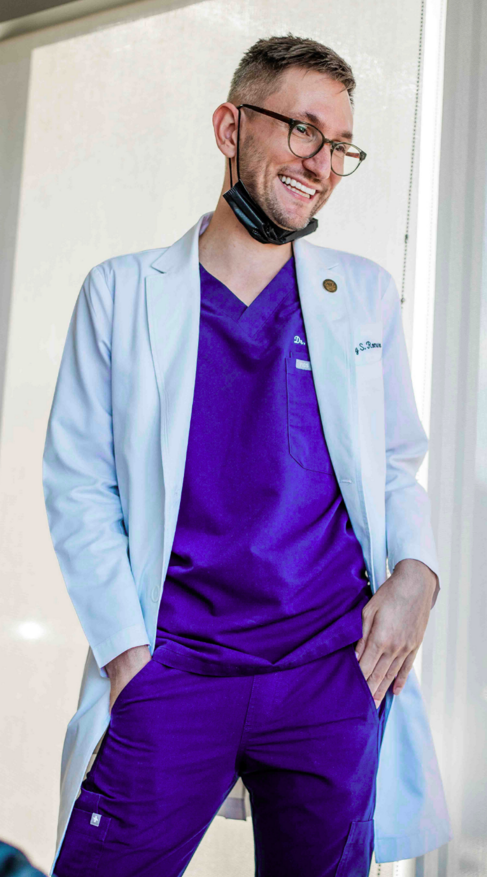 A male healthcare professional in purple scrubs and a white lab coat, smiling with hands in pockets, wearing glasses and a black face mask hanging below his chin, standing near a window.