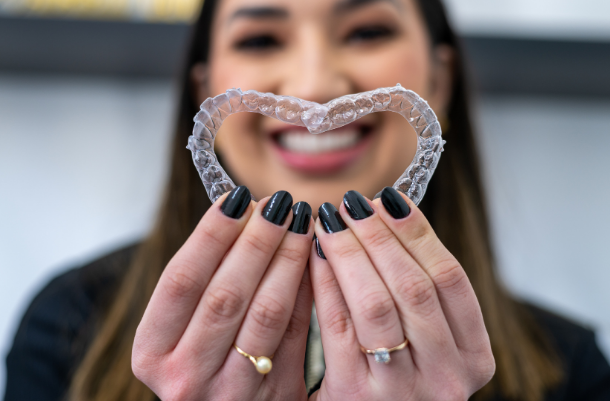 A woman smiling and holding a clear dental retainer in the shape of a heart in front of her face.