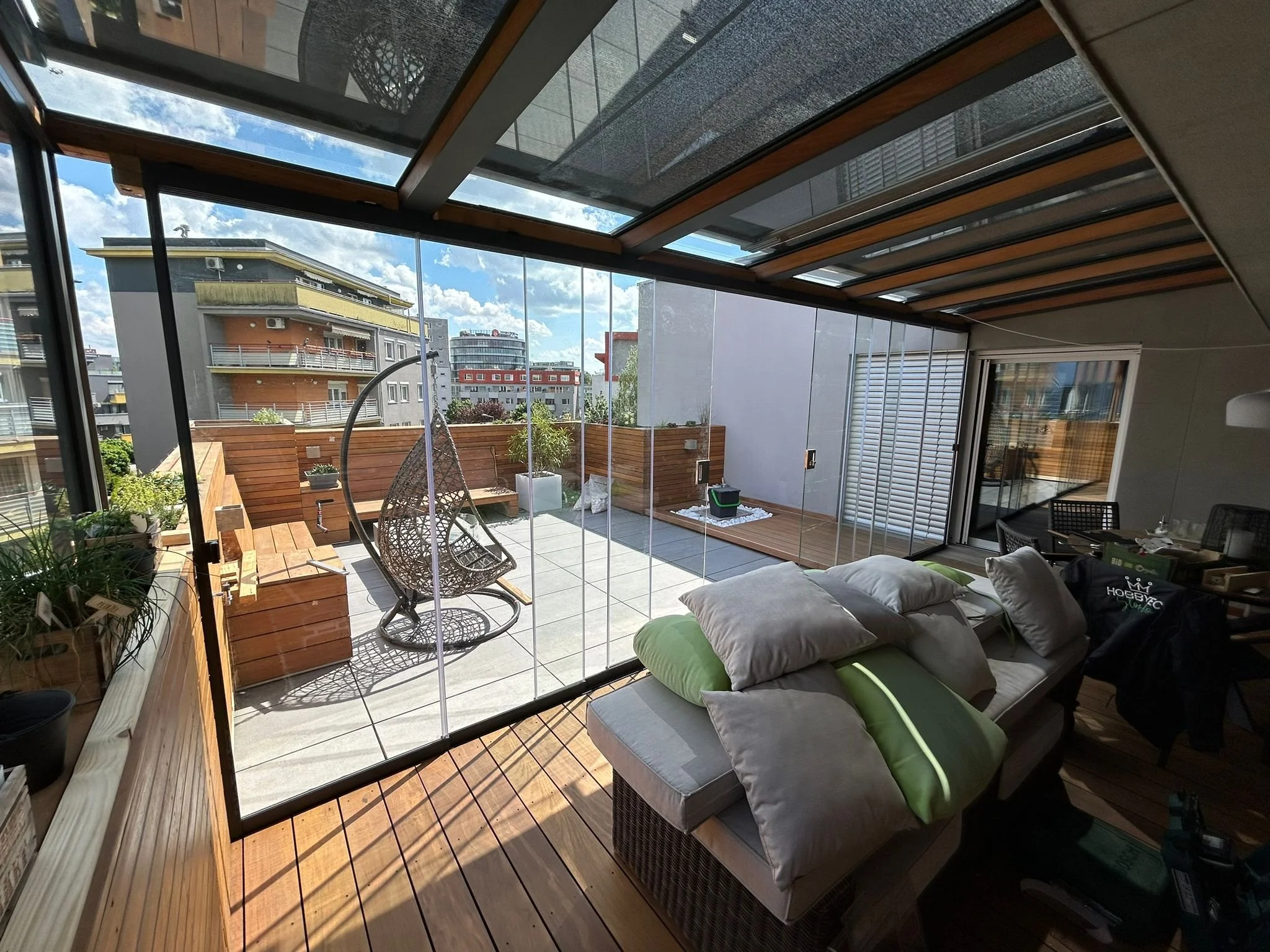 Modern tetőterasz és üvegterasz beépítés keret nélküli eltolható üvegfalakkal, üvegtetővel és elegáns fa burkolattal egy városi tetőkertben, pihenőbútorokkal és panorámás kilátással.