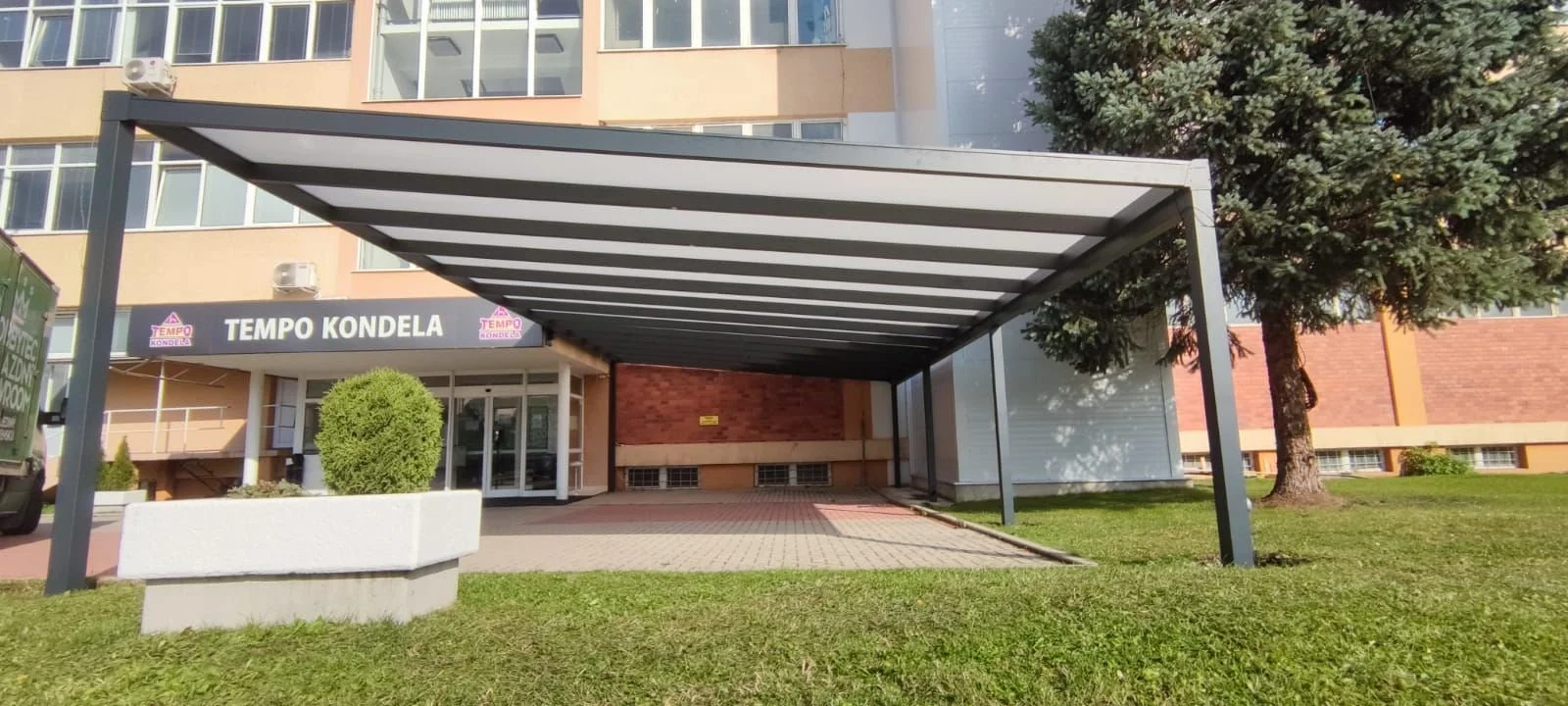 Bejárati előtető alumínium szerkezettel, fehér kaspós díszbokorral és zöld füves területtel - modern alumínium előtető és teraszfedés megoldás