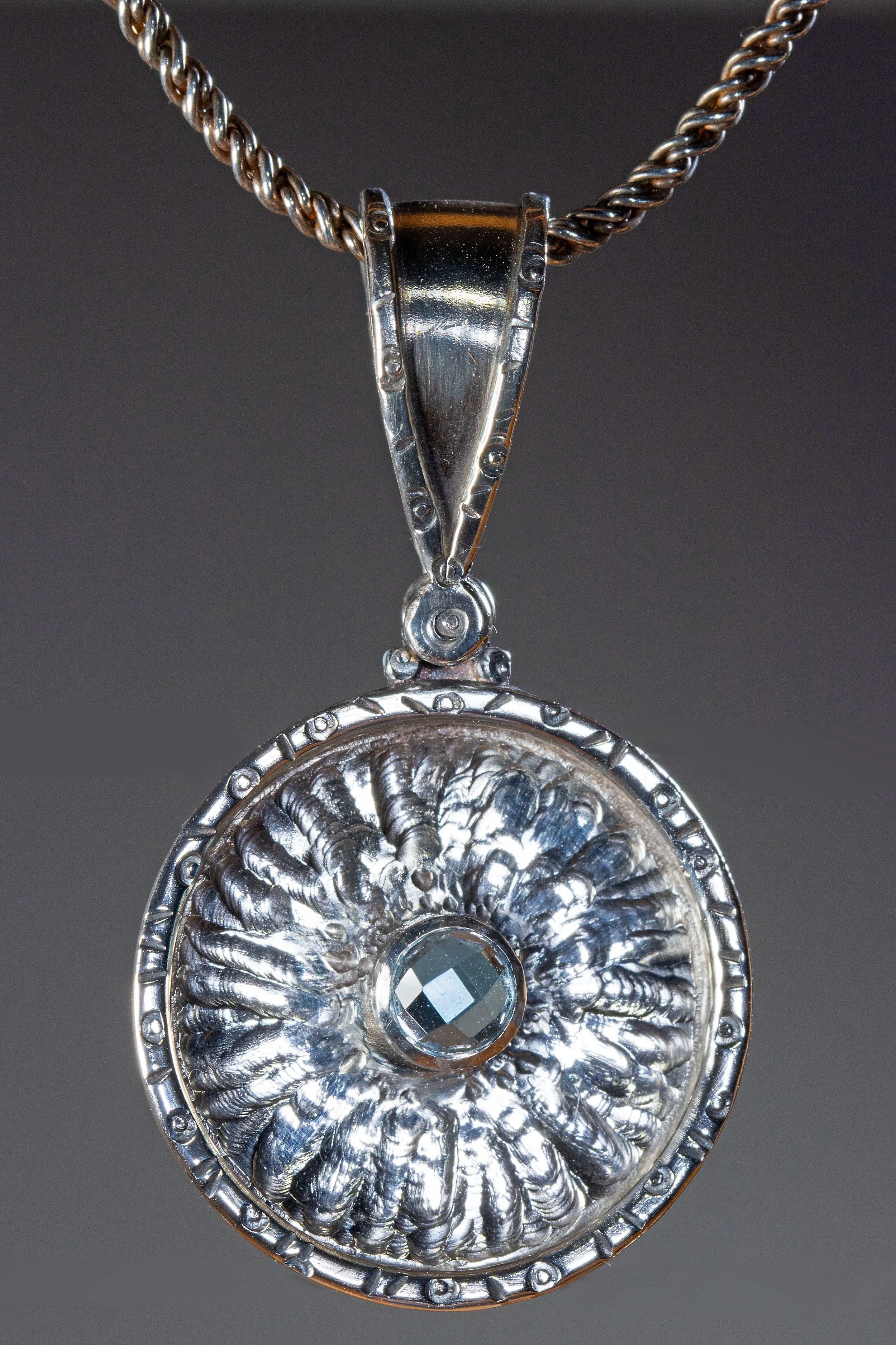 Close-up of a silver pendant necklace with a round, ornate, filigree design and a central faceted gemstone, hanging from a twisted silver chain against a dark background.