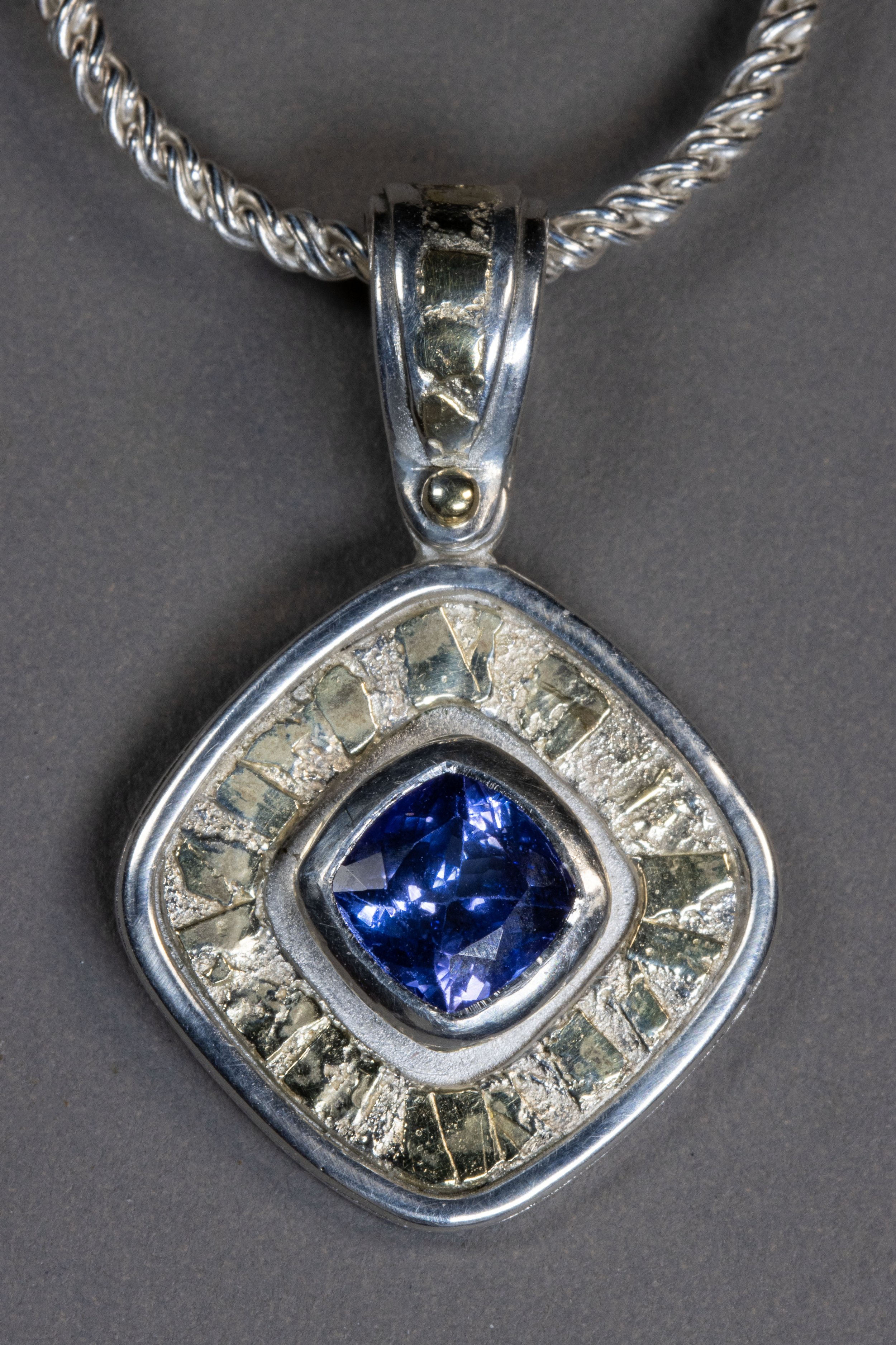 A close-up of a pendant featuring a large, deep blue gemstone encased in a silver setting with small, clear stones surrounding it, attached to a twisted silver chain.