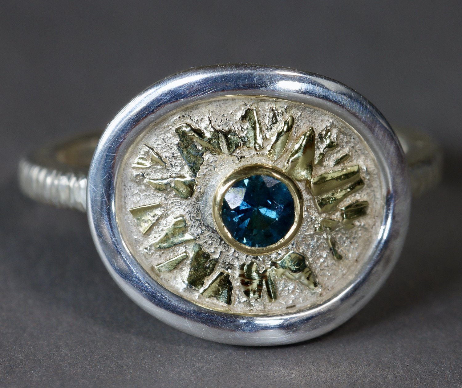 Close-up of a metallic ring with a central blue gemstone surrounded by sunburst-like metal detailing, against a dark background.