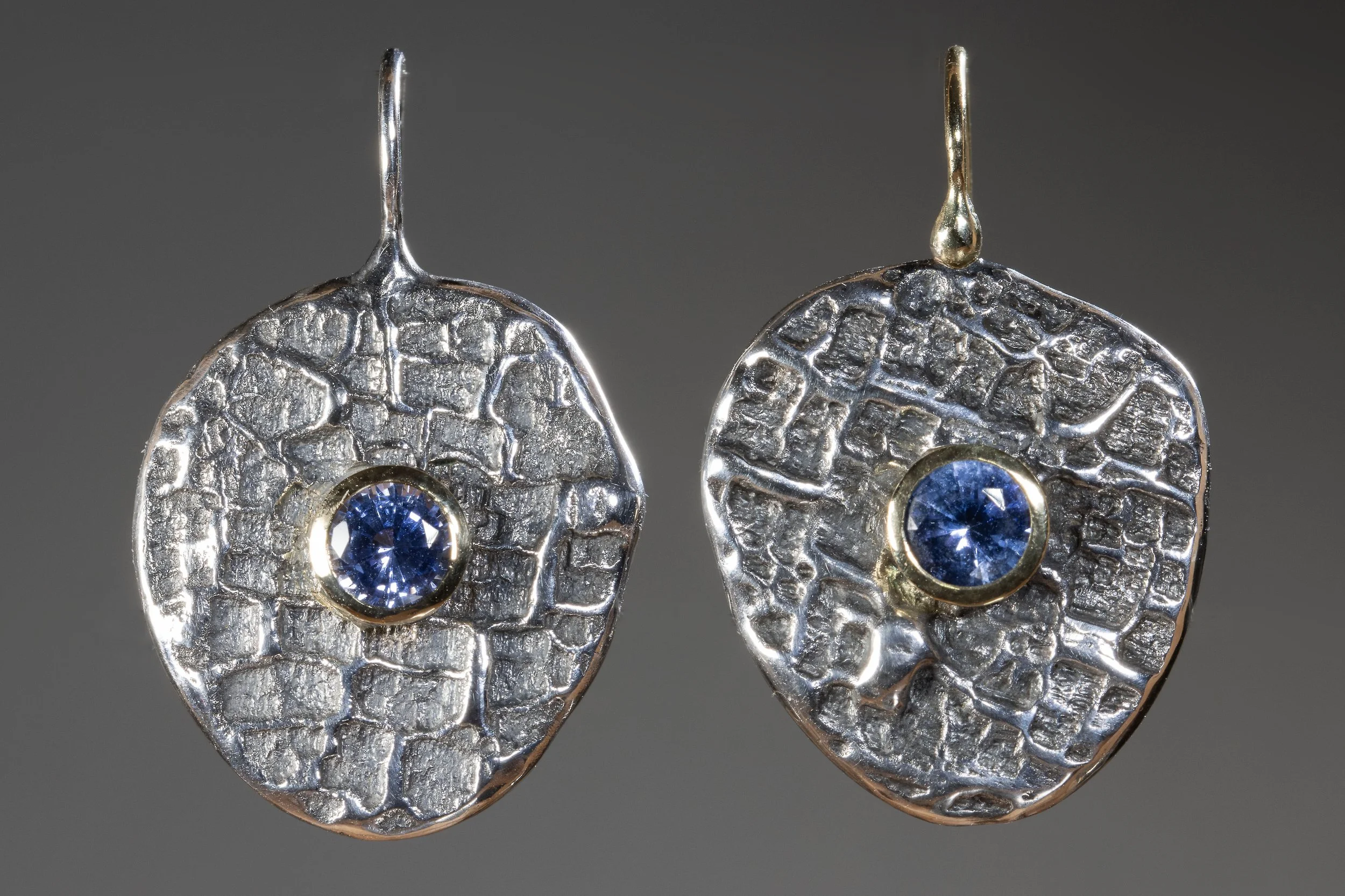 Close-up of silver, textured earrings with blue gemstones in the center, hung against a plain dark background.