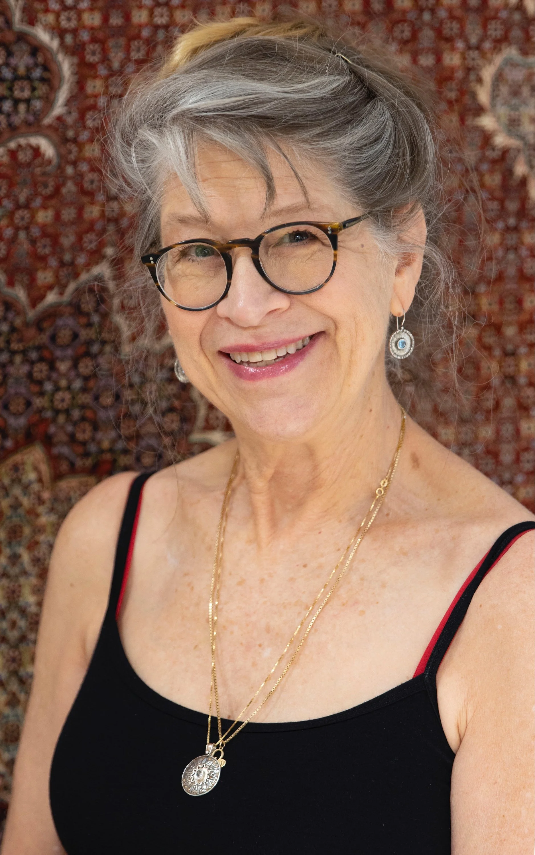 Cory Lawson with gray hair, wearing glasses, earrings, and layered gold necklaces, dressed in a black tank top with red trim, standing against a patterned rug background.