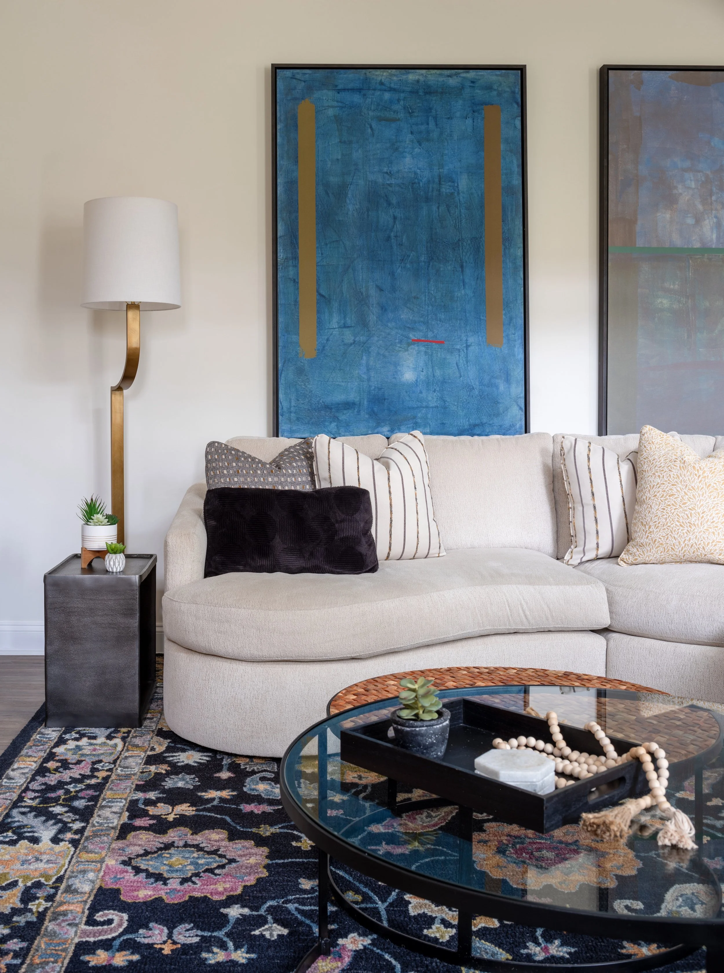 Living room designed by KD Designs with a beige sofa, decorative pillows, a glass coffee table with a tray, a blue abstract painting on the wall, a side table with plants, and a tall floor lamp.