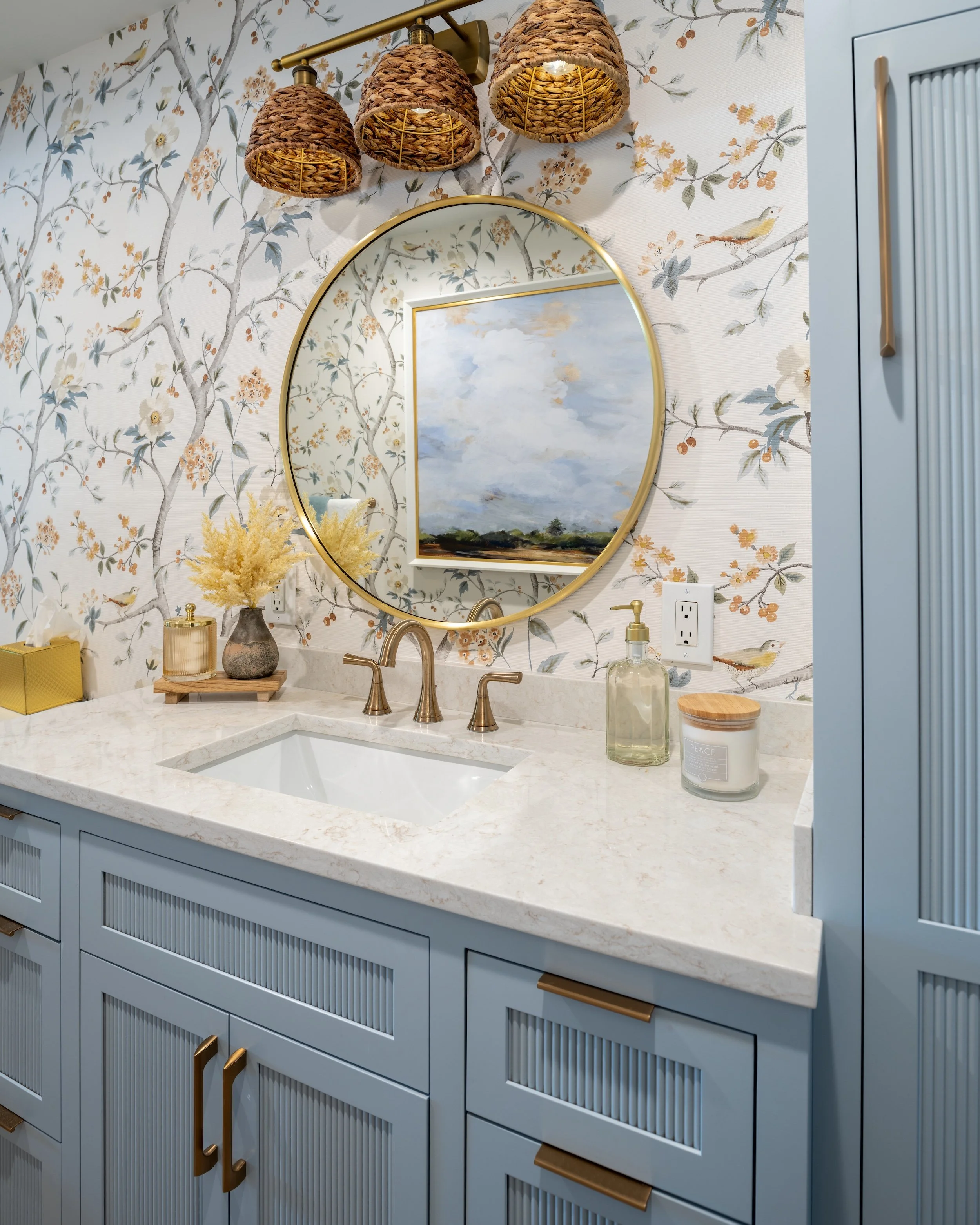 Bathroom by KD Designs with blue cabinetry, marble countertop, round mirror, floral wallpaper, and woven light fixtures.