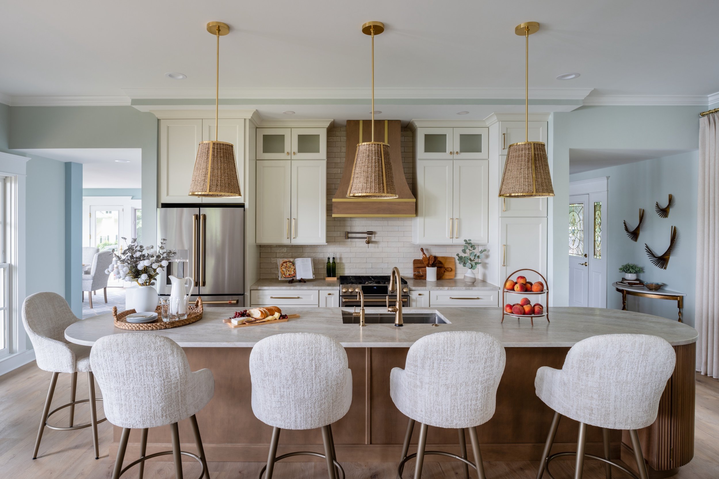 KD Designs kitchen with white cabinets, stainless steel refrigerator, and a large island with white cushioned stools. There are three pendant lights with wicker shades hanging above the island.