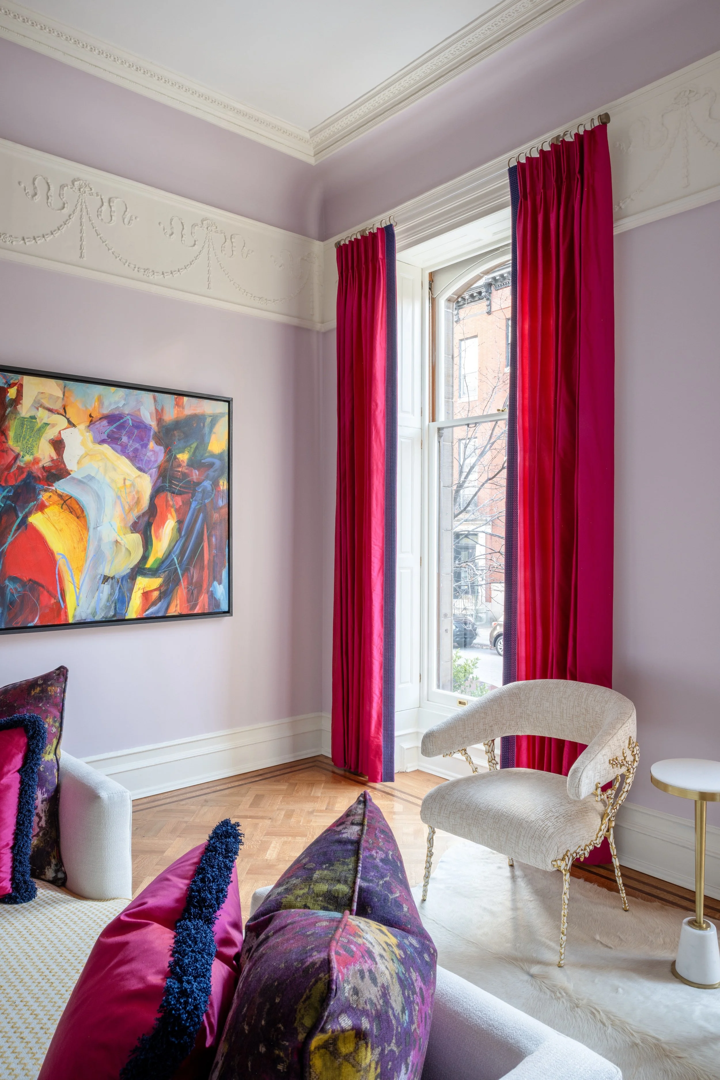A colorful living room designed by KD Designs with light purple walls, red curtains, a window showing buildings outside, a beige copy chair, and a sofa with purple and pink cushions.
