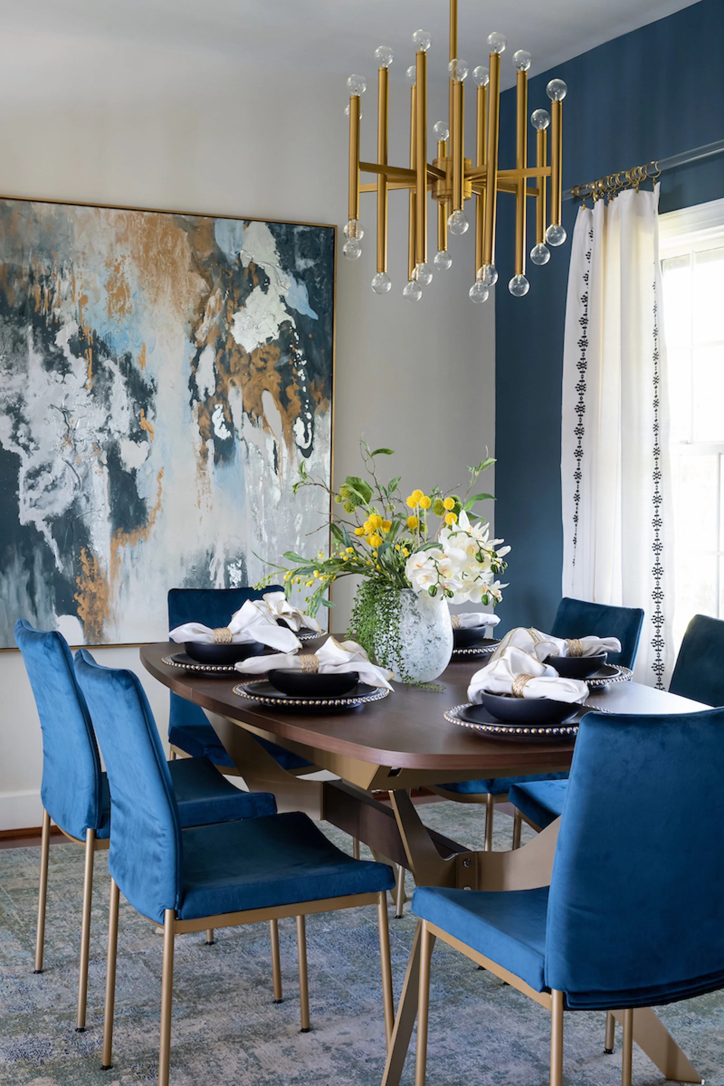 A dining room designed by KD Designs with a dark wood table, six blue velvet chairs with gold legs, a large abstract painting on the wall, a modern gold chandelier hanging above, and a white curtain on the window.