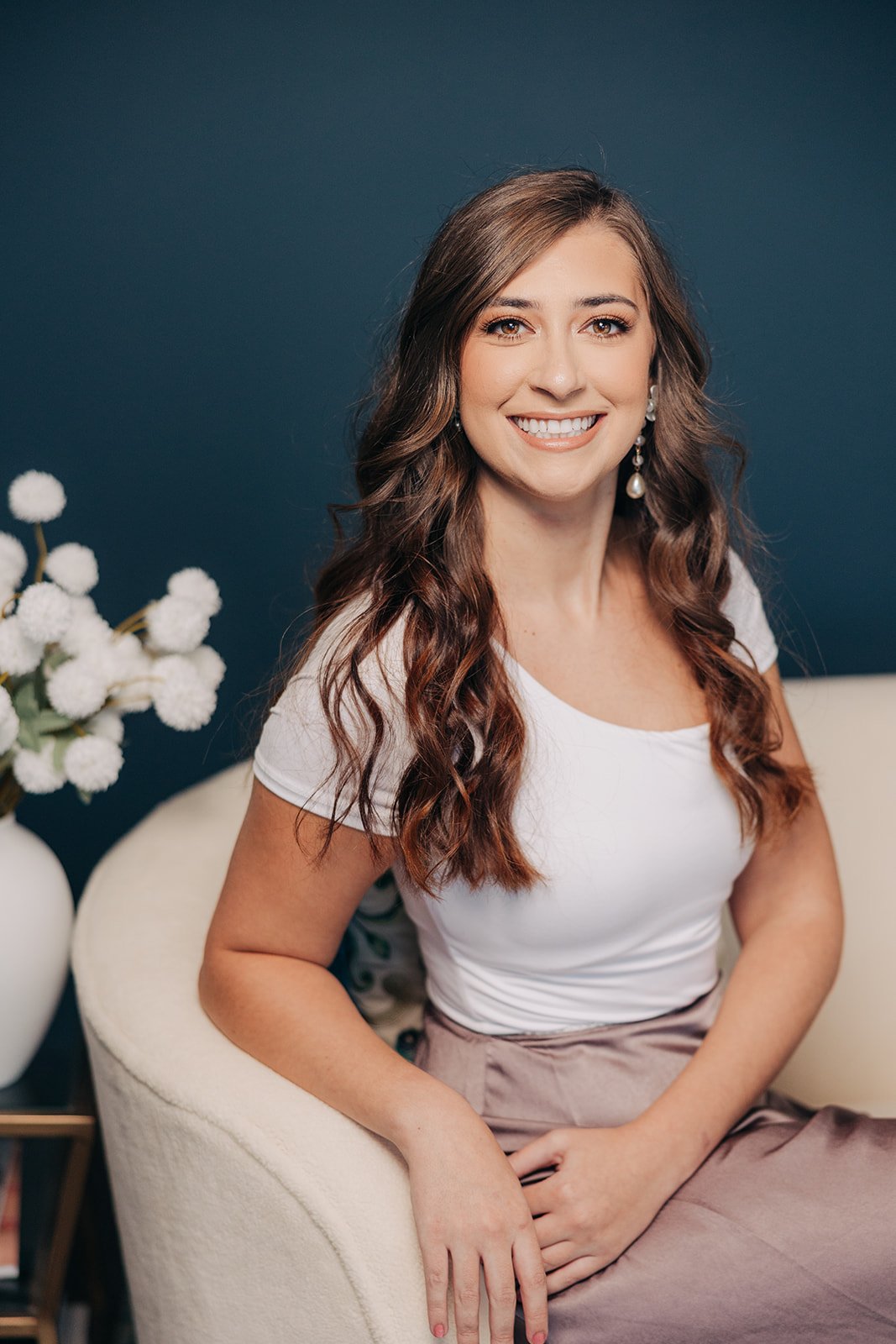 Associate designer Bethany Finn seated in a chair, smiling, wearing a white top and neutral skirt against a dark blue backdrop, representing KD Designs, an interior design firm based in Anne Arundel County, MD