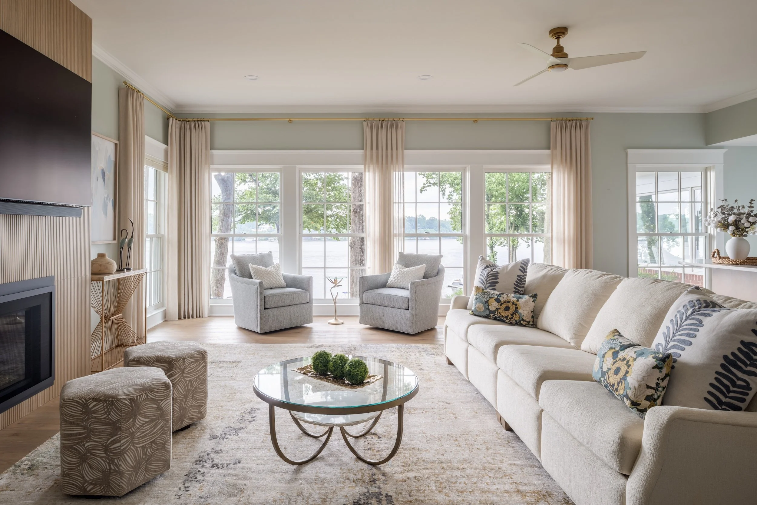 Bright living room designed by KD Design with large windows overlooking a lake, with beige sofa, two gray armchairs, and a glass coffee table with decorative green spheres, and a fireplace on the left.