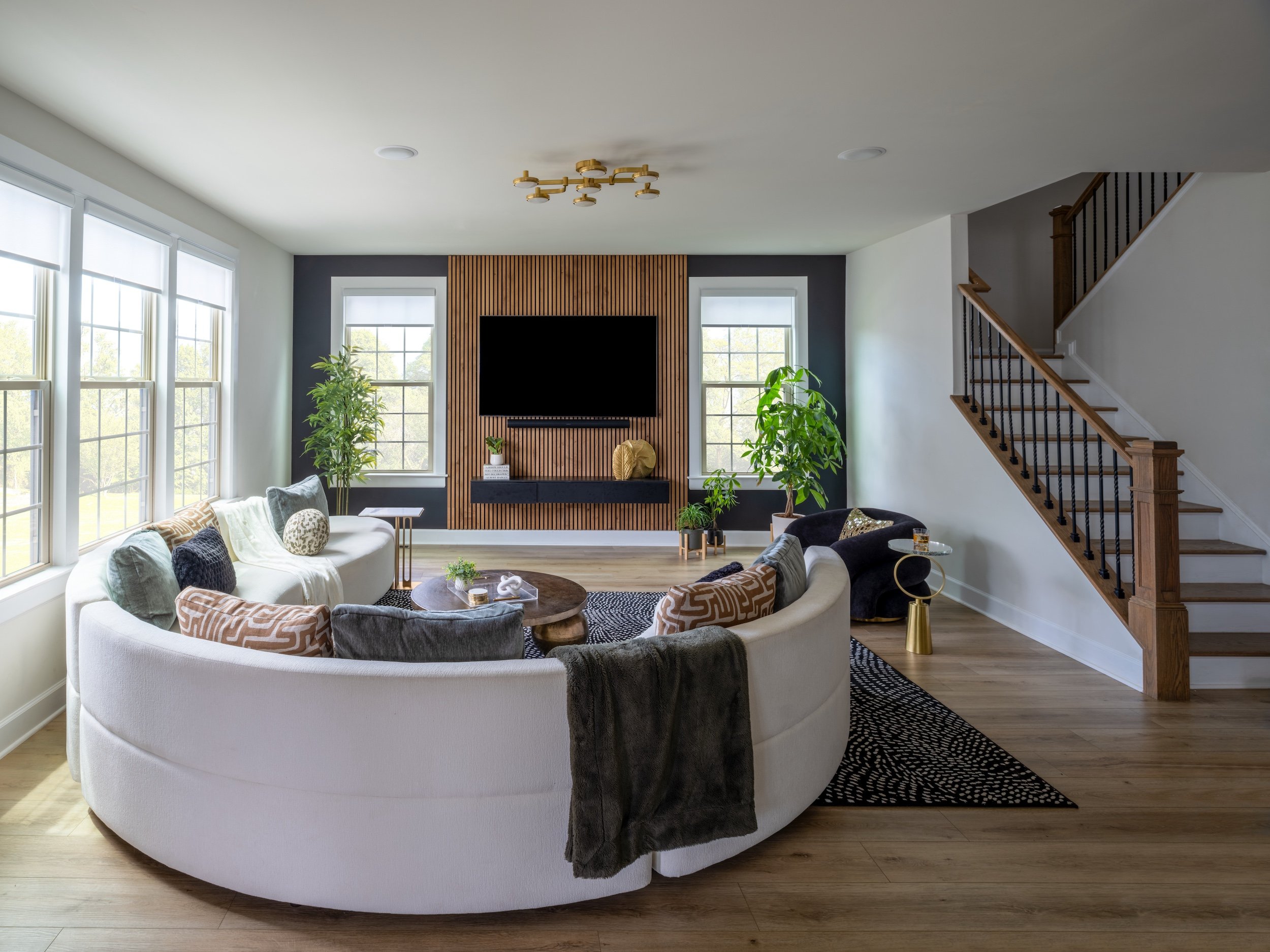 Living room designed by KD Designs with a curved white sofa, black and beige pillows, and a dark wood coffee table, near large windows with white blinds, a flat-screen TV on a wooden panel wall and staircase with wooden handrail.