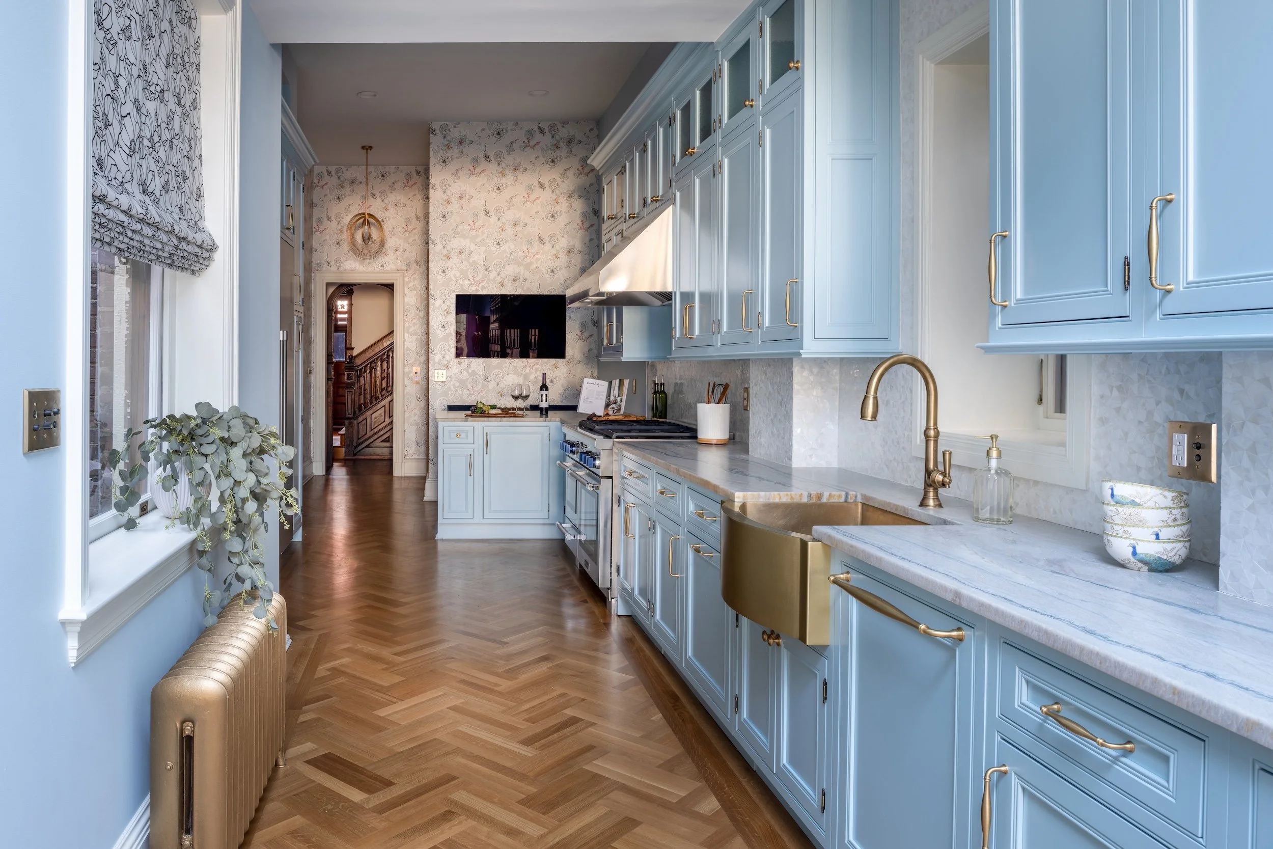 A spacious kitchen designed by KD Designs with blue cabinets, gold hardware, marble countertops, hardwood floor, a window with patterned Roman shade, and a golden radiator. At the end, a hallway with wallpaper and a staircase is visible.