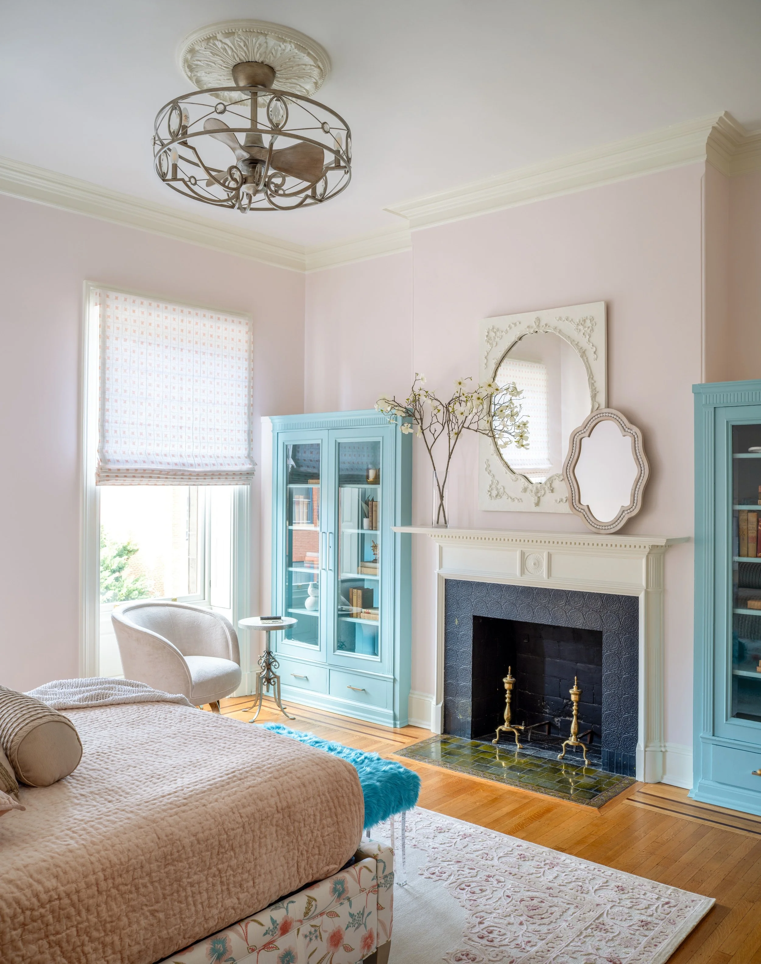 A living room by KD Designs with a beige quilted bed, a white and floral ottoman, a cream armchair, a blue display cabinet, a white fireplace with decorative items and a mirror above it, hardwood flooring, a patterned area rug, and a ceiling fan.
