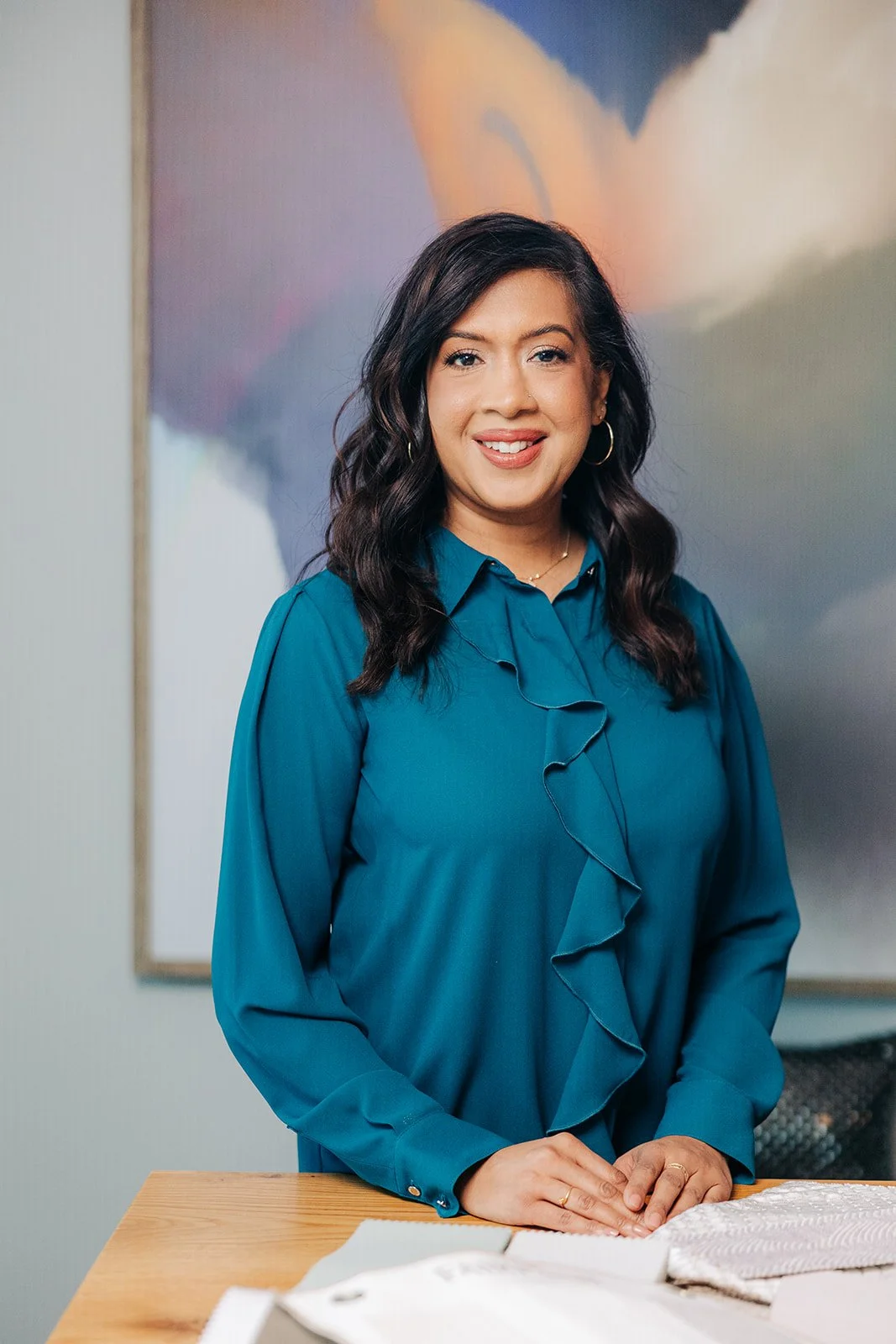 KD Designs Design Coordinator Jeehan Kashim wearing a teal blouse, smiling, standing behind a wooden table with fabric swatches.