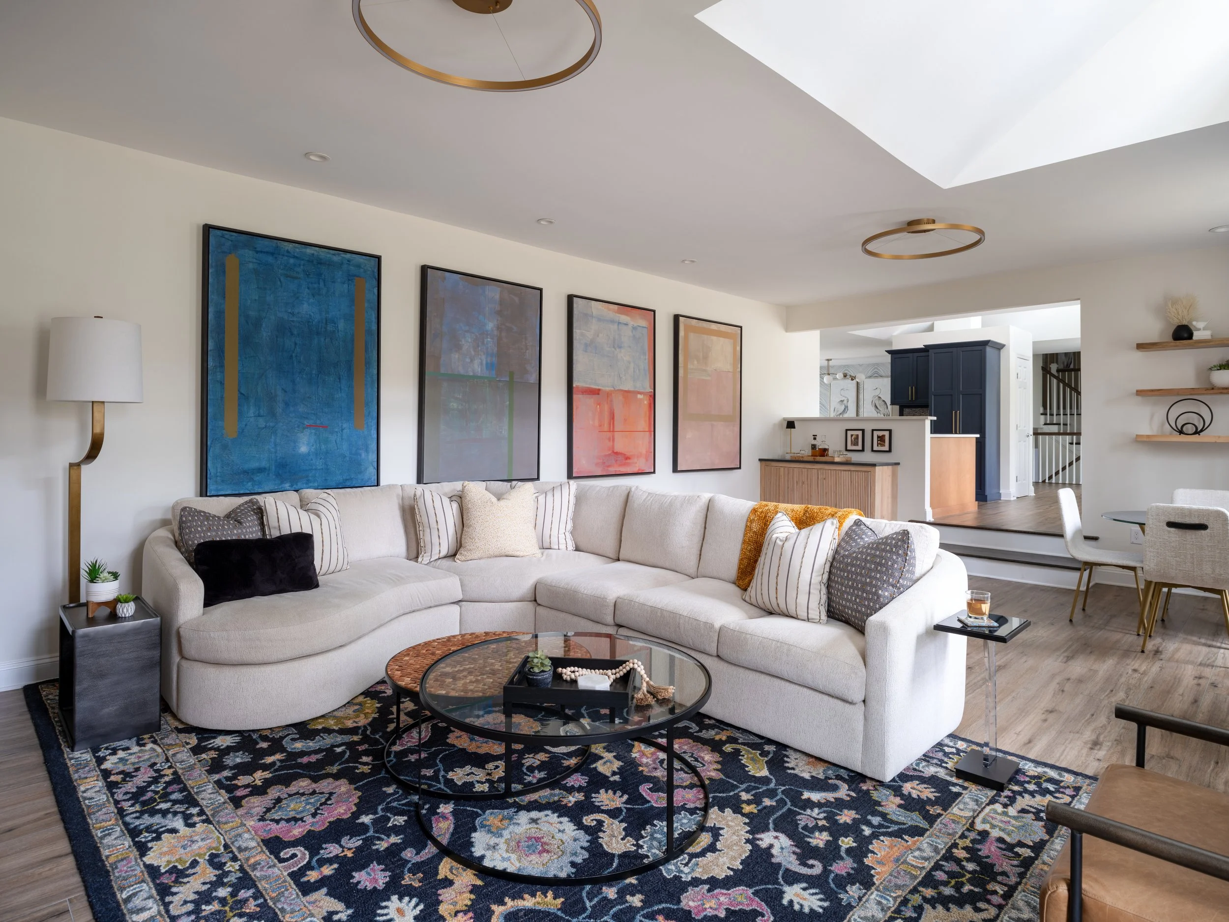 Living room designed by KD Designs with a beige sectional sofa, abstract wall art, a black coffee table, a black and beige rug, a white floor lamp, and decorative pillows; open concept with glimpses of kitchen and dining area.