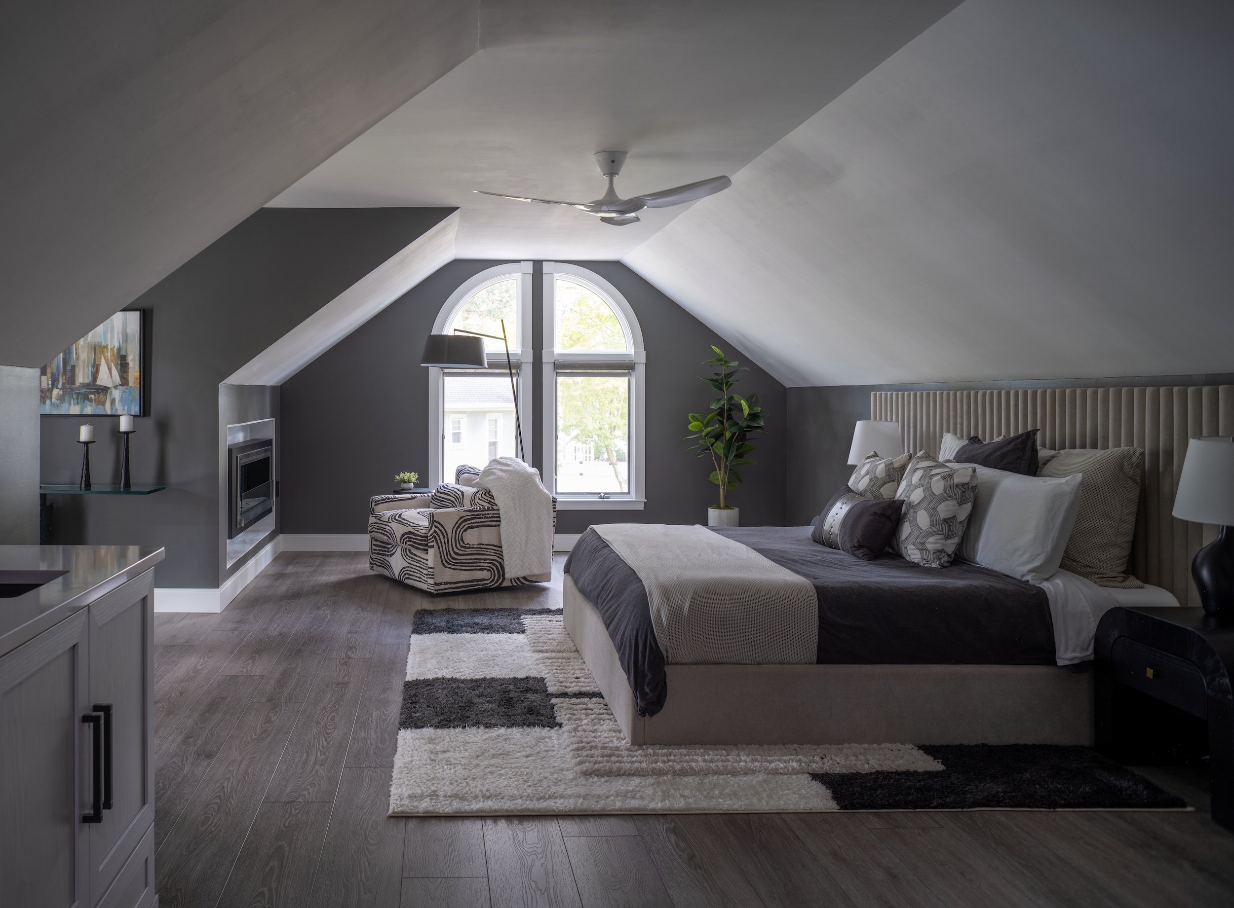 Modern bedroom designed by KD Designs with a sloped ceiling, large window, bed with multiple pillows, a side table with a lamp, a patterned armchair, and a potted plant.