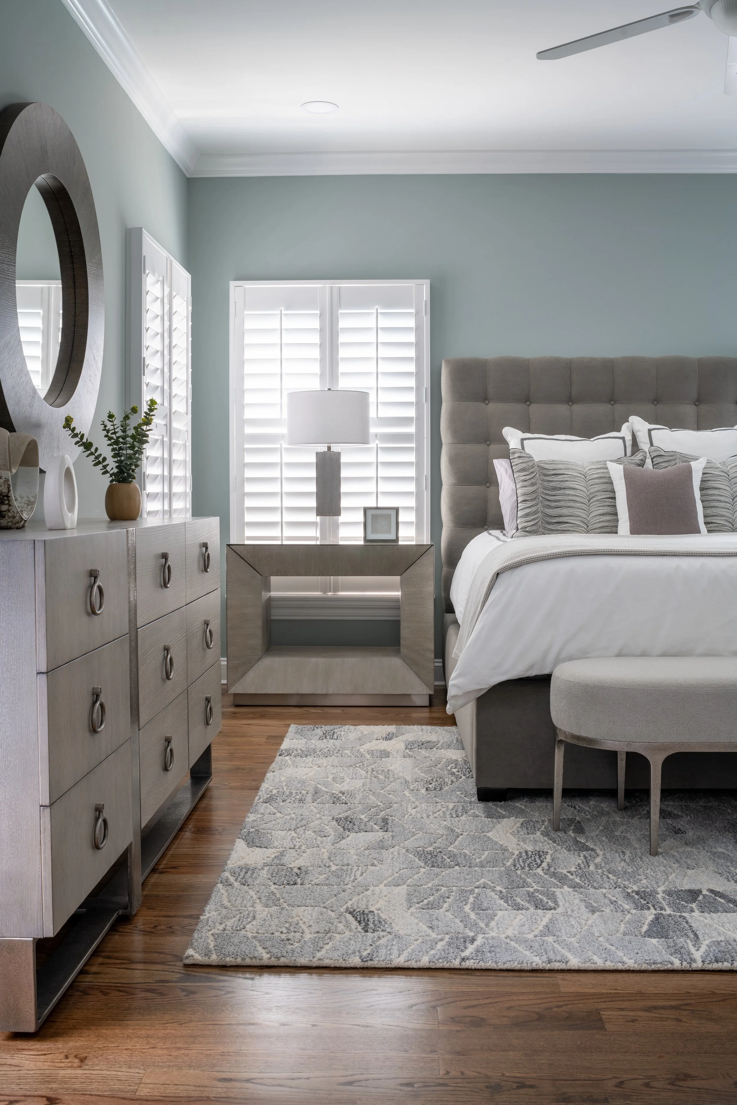 A modern bedroom designed by KD Designs with a large upholstered bed, multiple decorative pillows, a bedside table with a lamp, a dresser with decor items, and a rug on wooden flooring.