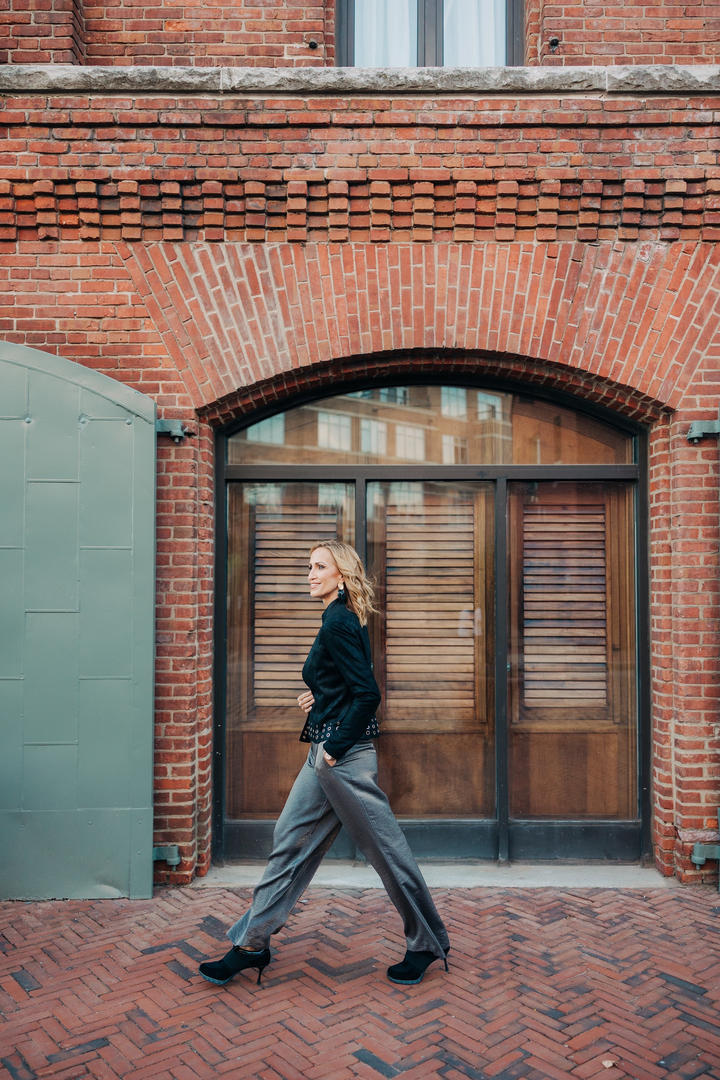 KD Designs founder and principal Krista Shugars in a black blazer and gray plaid pants walking past a red brick building with a large window and wooden shutters in Baltimore.