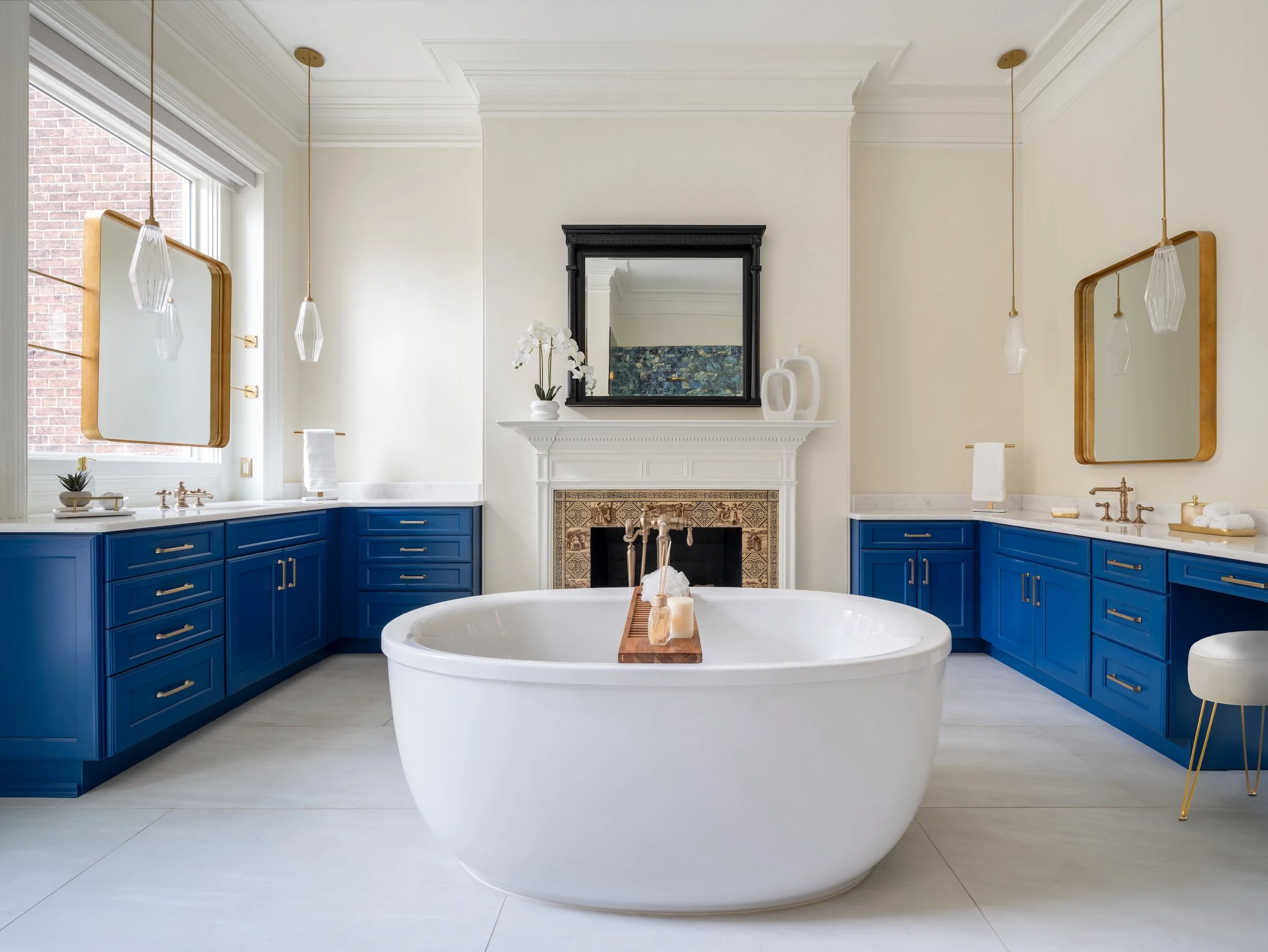 Modern bathroom designed by KD Designs with a white oval bathtub, blue cabinets, and gold fixtures, a fireplace, and a large window.