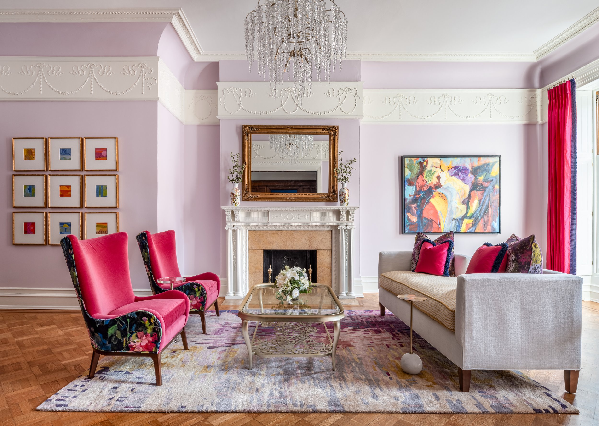 A living room by KD Designs with pink walls and a white fireplace, decorated with a gold-framed mirror and vases on the mantle. There are two red-columnwing chairs with floral upholstery, a white sofa with pink pillows, and a glass-top coffee table.