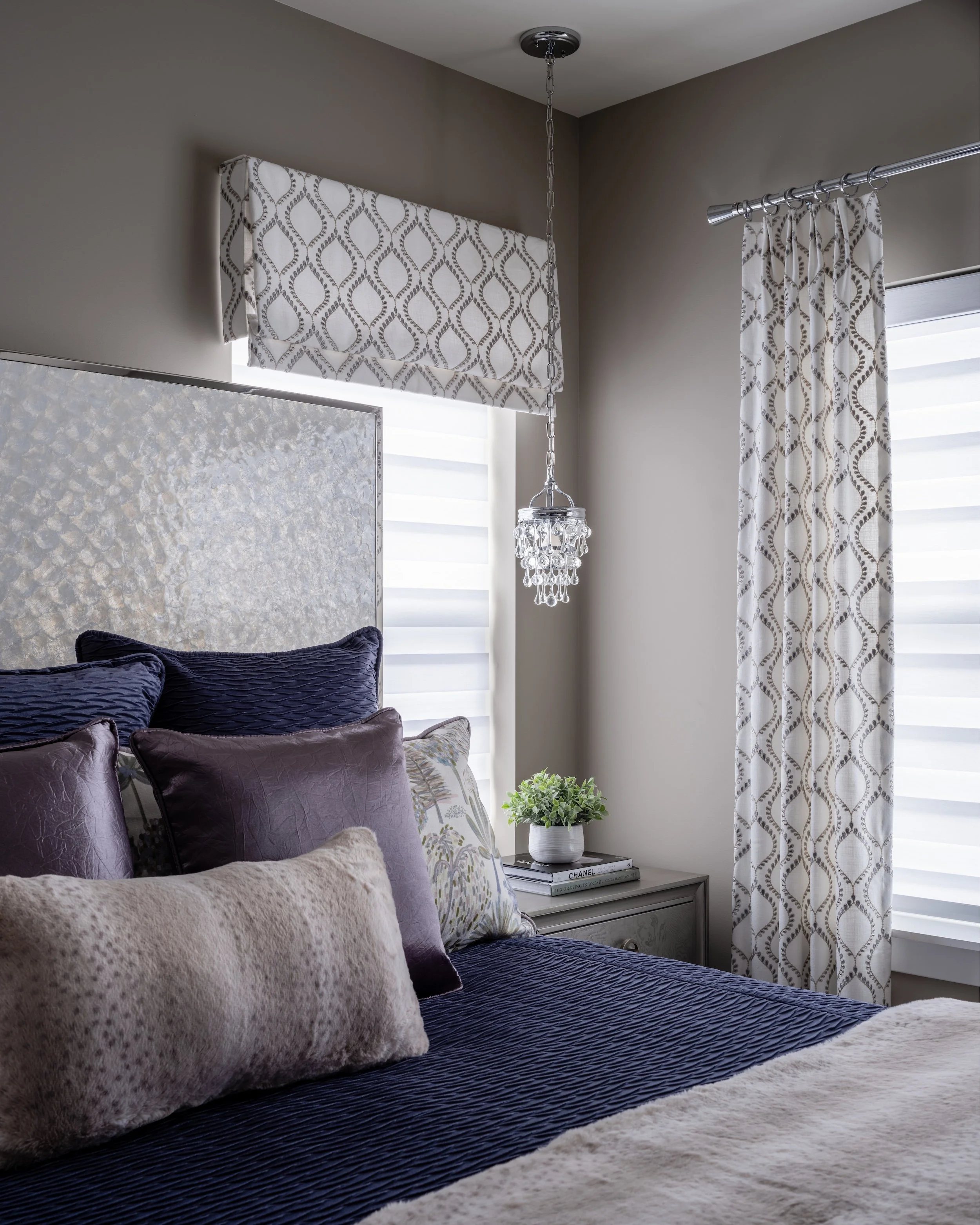 A bedroom designed by KD Designs with a bed featuring dark purple and beige pillows, a gray nightstand with books and a potted plant, lace curtains, a valance, and a small chandelier hanging from the ceiling.