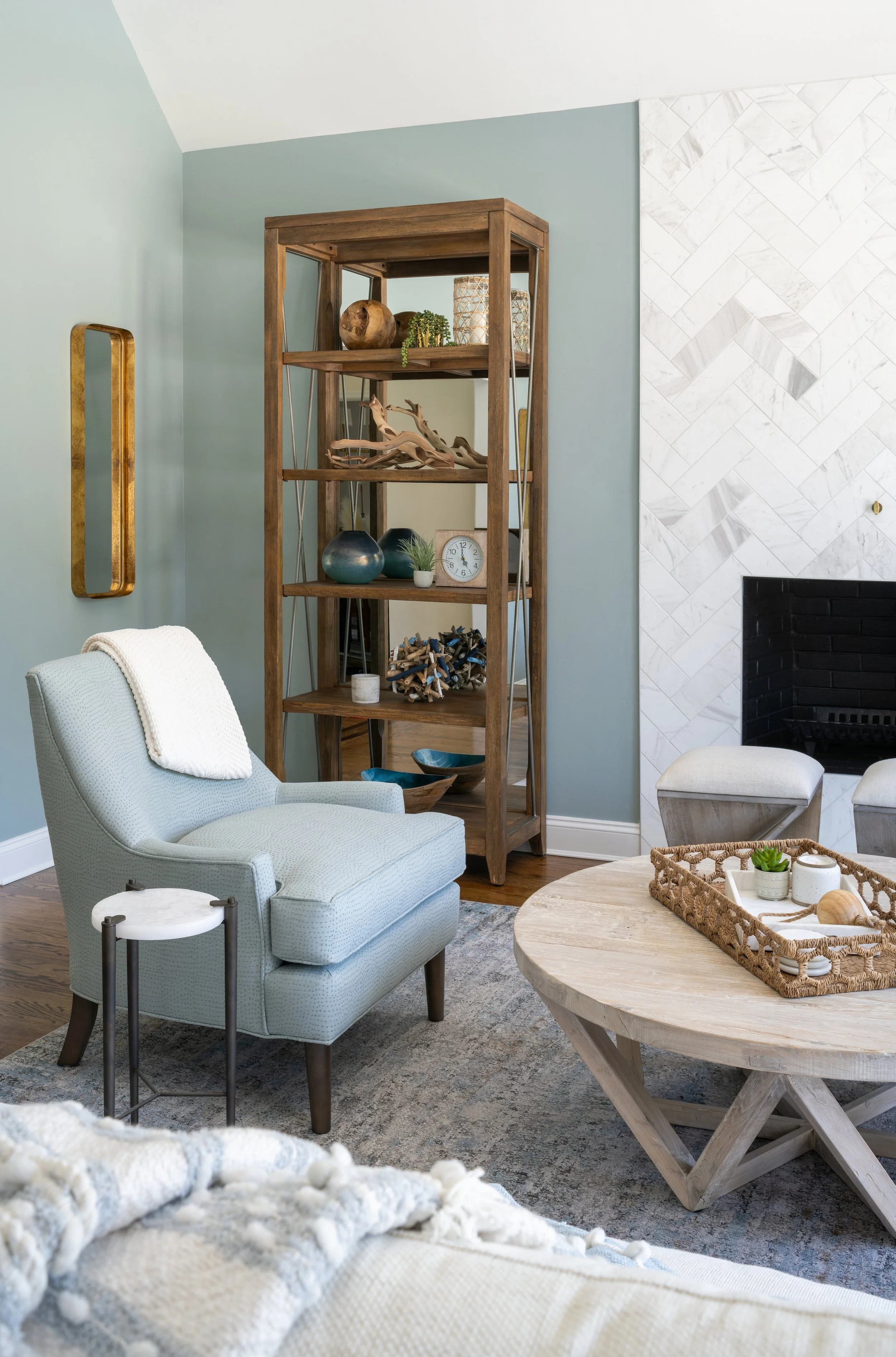 Coastal-inspired living room designed by KD Designs in Annapolis, MD, featuring a soft blue upholstered armchair, round wood coffee table, styled open shelving, and a marble fireplace against pale blue walls.