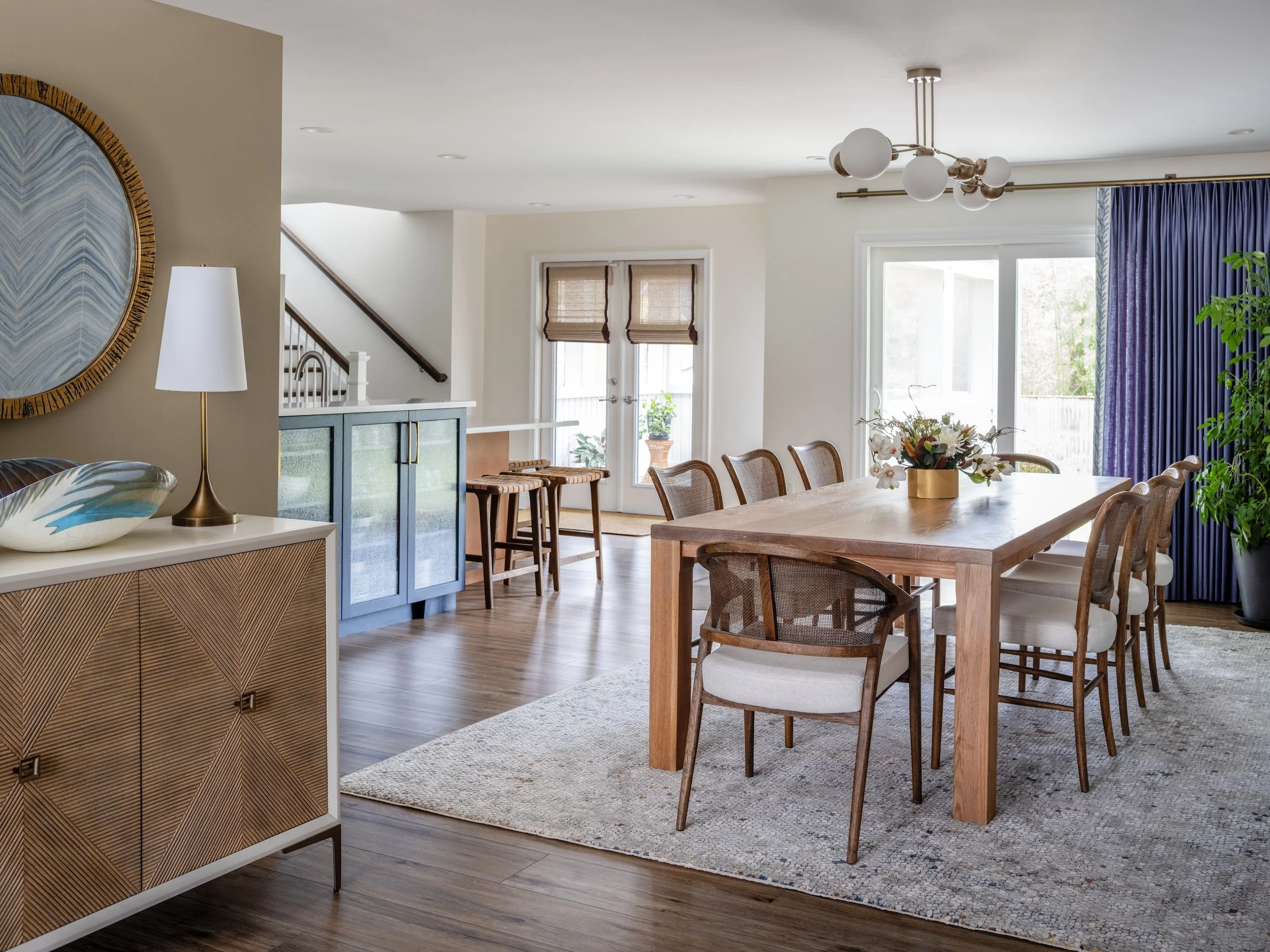Modern dining room designed by KD Designs with a wooden table, eight chairs, a floral centerpiece, and a large window with purple curtains.
