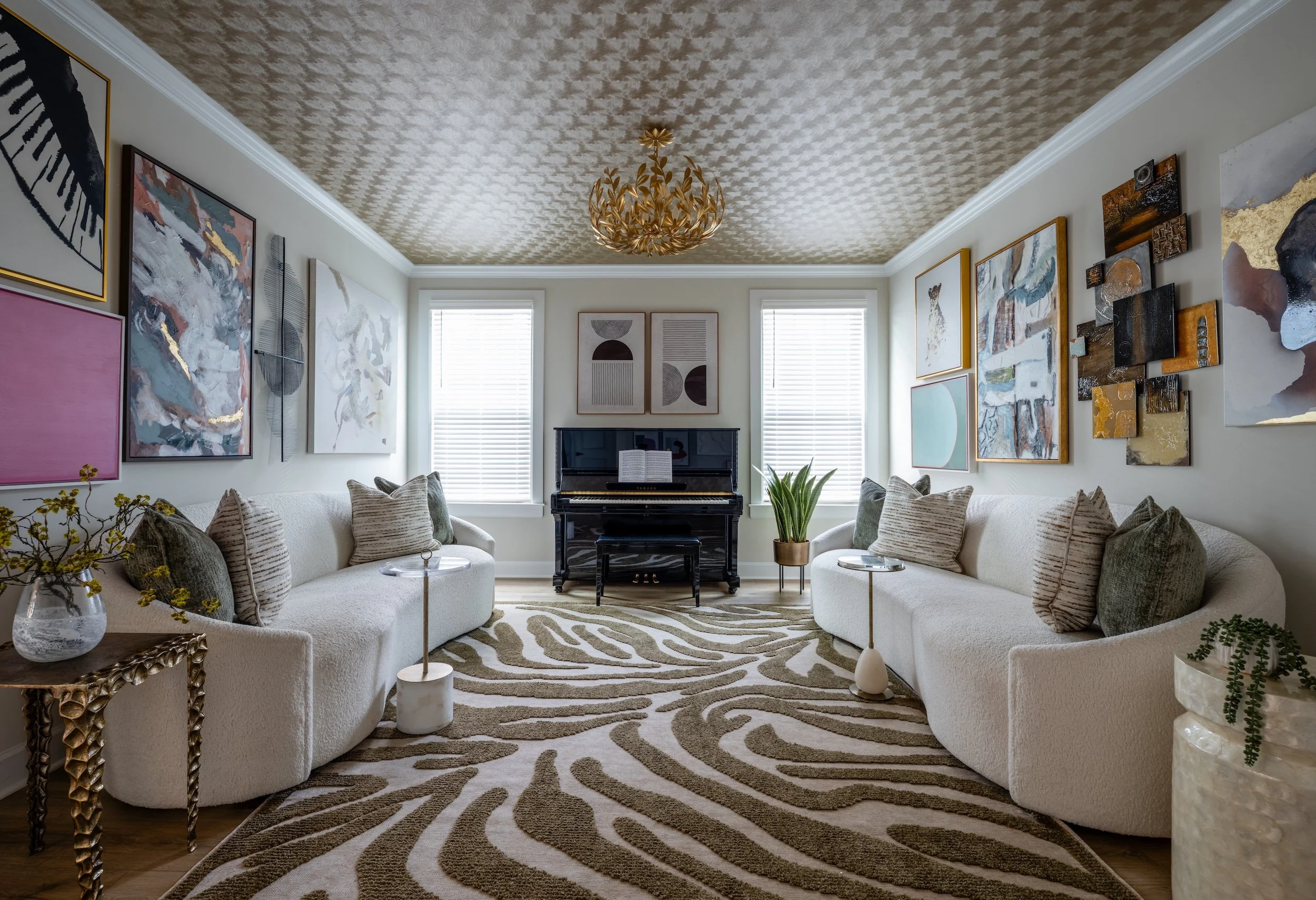 Living room with two white curved sofas, abstract wall art, a black piano, a patterned carpet, and gold accents, with windows and a gold chandelier.