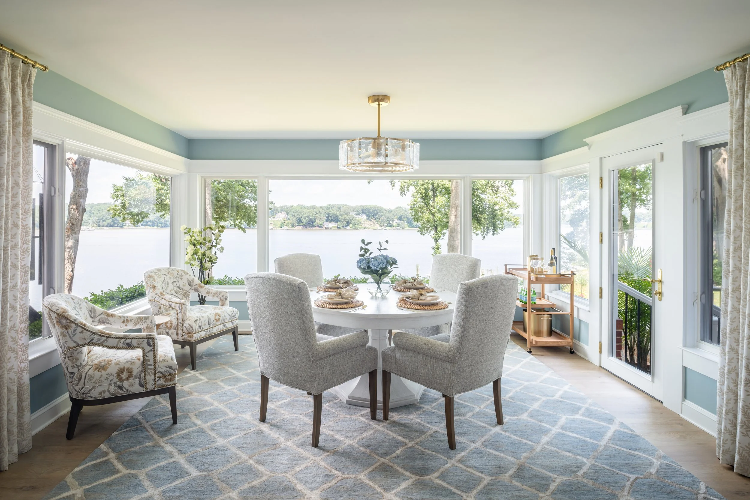 Bright dining room by KD Designs with large windows overlooking a body of water and trees, featuring a round table set for four with a vase of flowers, surrounded by upholstered chairs, and a light hanging fixture, with a door leading outside.