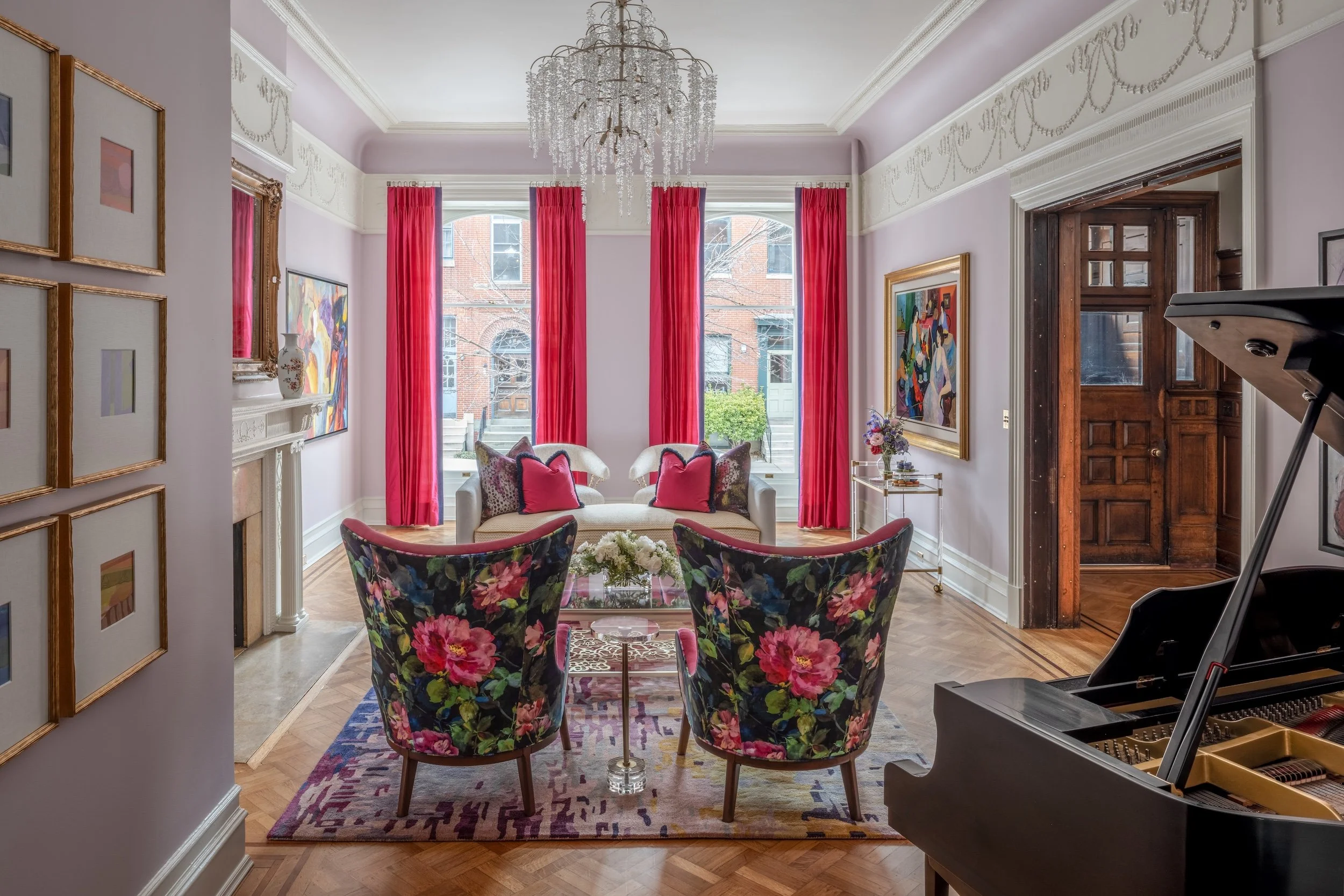 Living room of a home designed by KD Designs with a grand chandelier, two large windows with pink curtains, colorful artwork, floral armchairs, and a black grand piano.