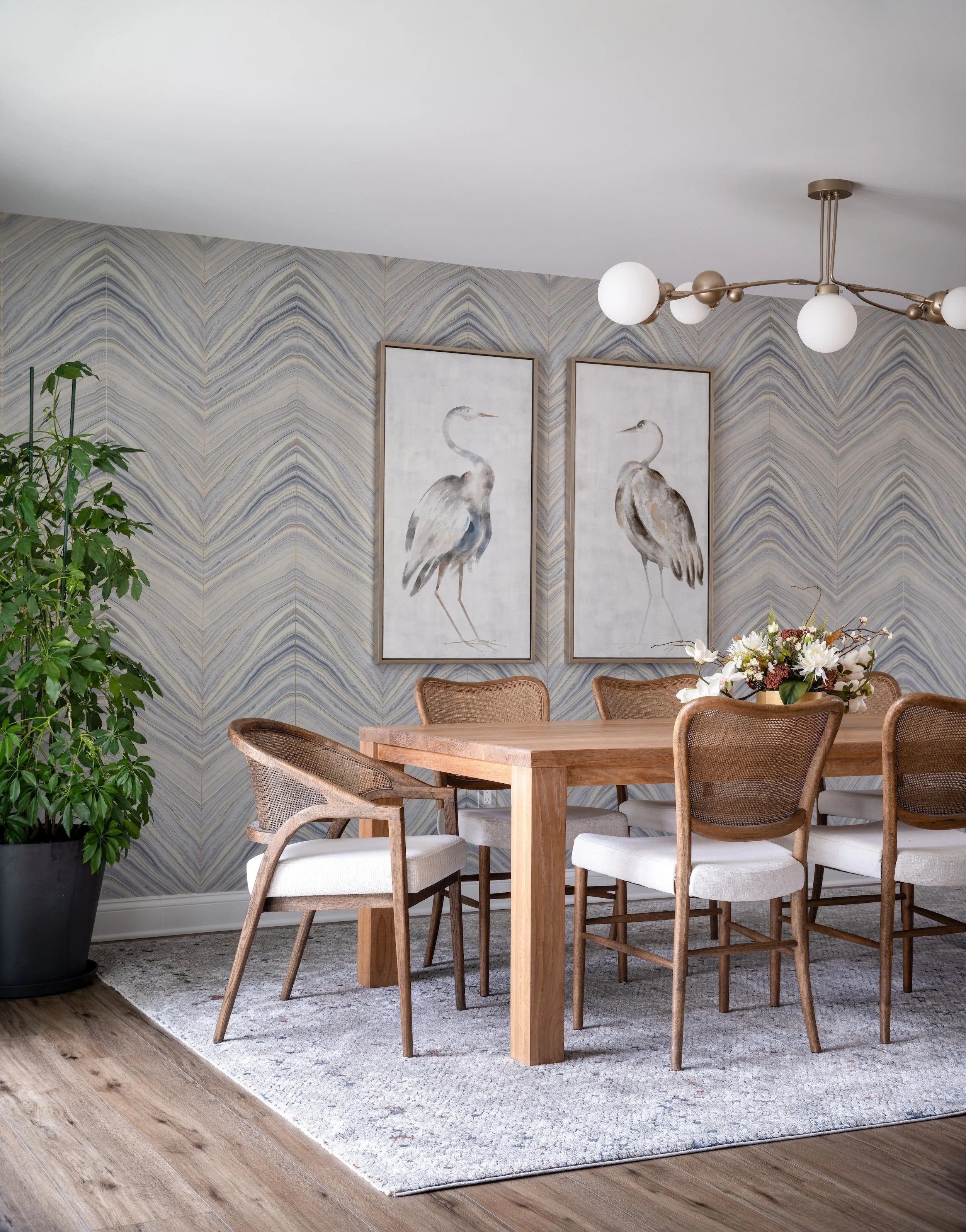 Dining room designed by KD Designs with wooden table, white and brown upholstered chairs, two framed artwork of herons on a textured wallpapered wall, a modern chandelier with globe lights, and a floral centerpiece on the table.