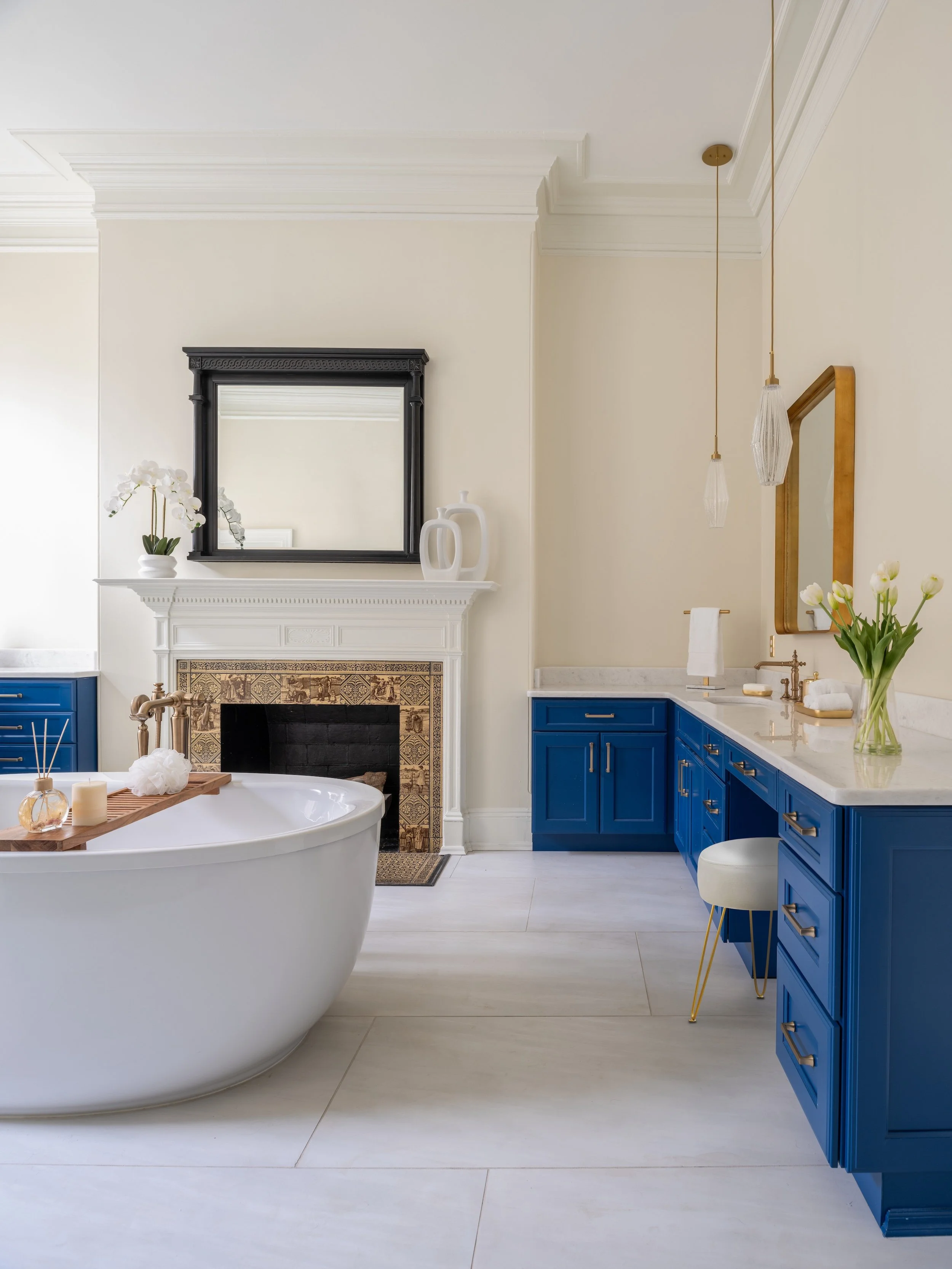 Modern bathroom designed by KD Designs with a white bathtub on the left, a fireplace with decorative tiles, blue cabinets, a marble countertop, and a large mirror with a gold frame on the right.