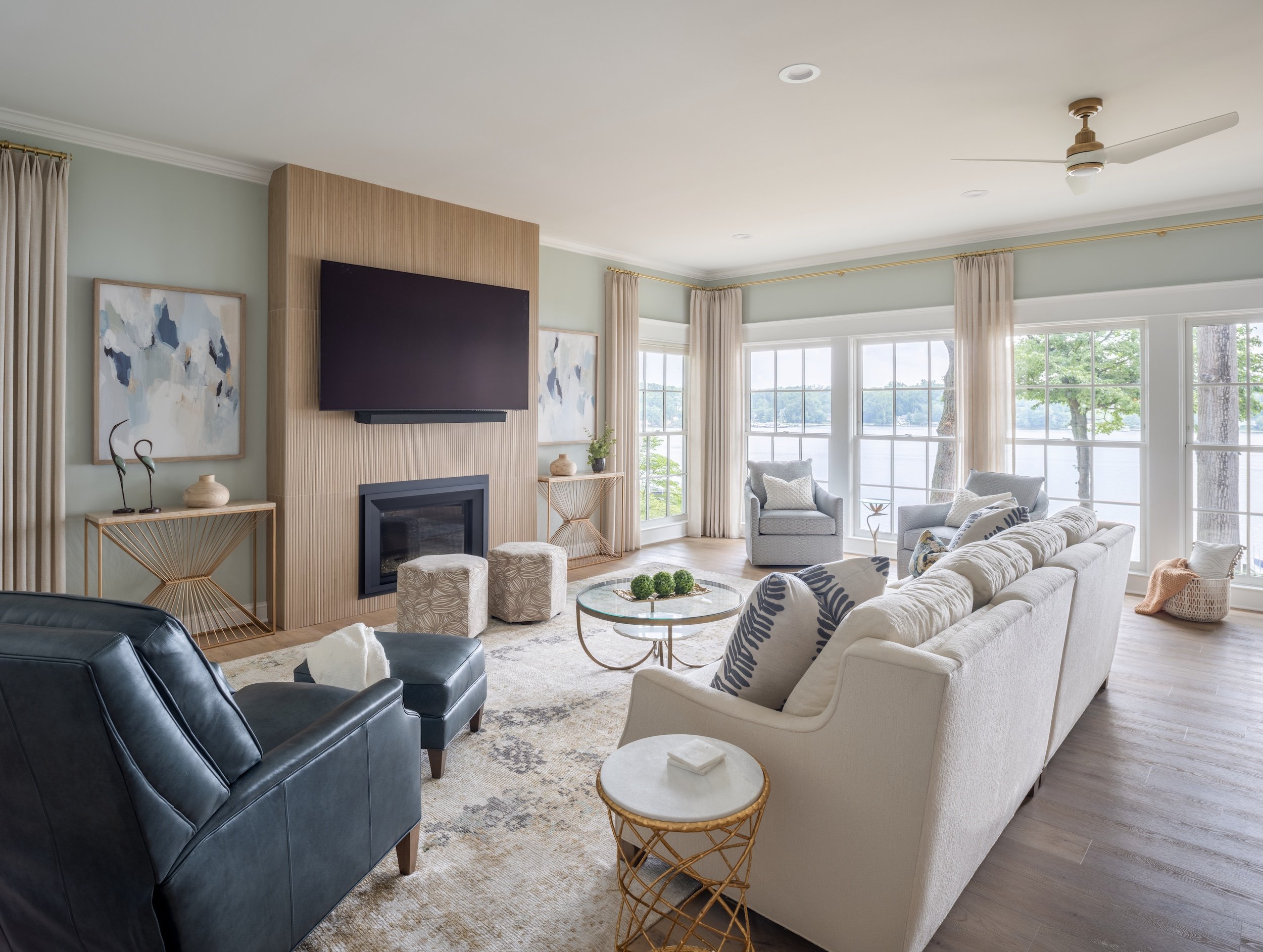 A living room designed by KD Designs with large windows overlooking a lake, featuring a cream sofa, armchairs, a black leather chair, and modern decor.