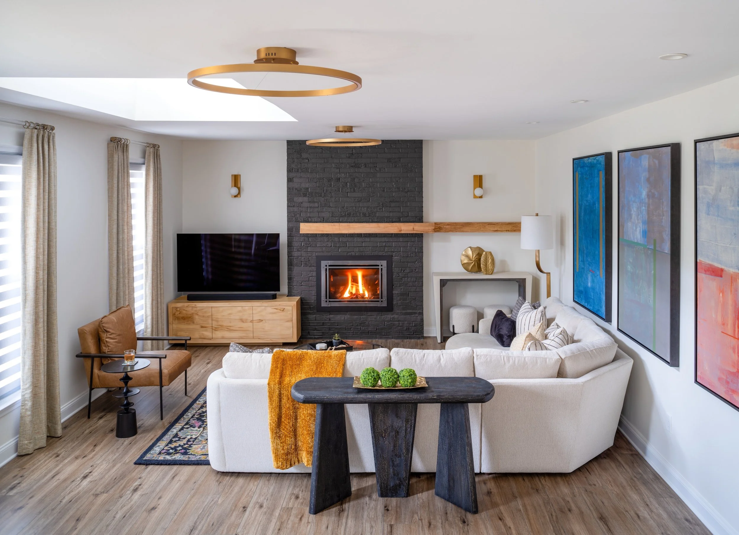 Living room by KD Designs with white sectional sofa, black wood table, tan armchair, TV on wooden stand, black brick fireplace with fire, abstract art on walls, wooden floor, and ceiling lights.