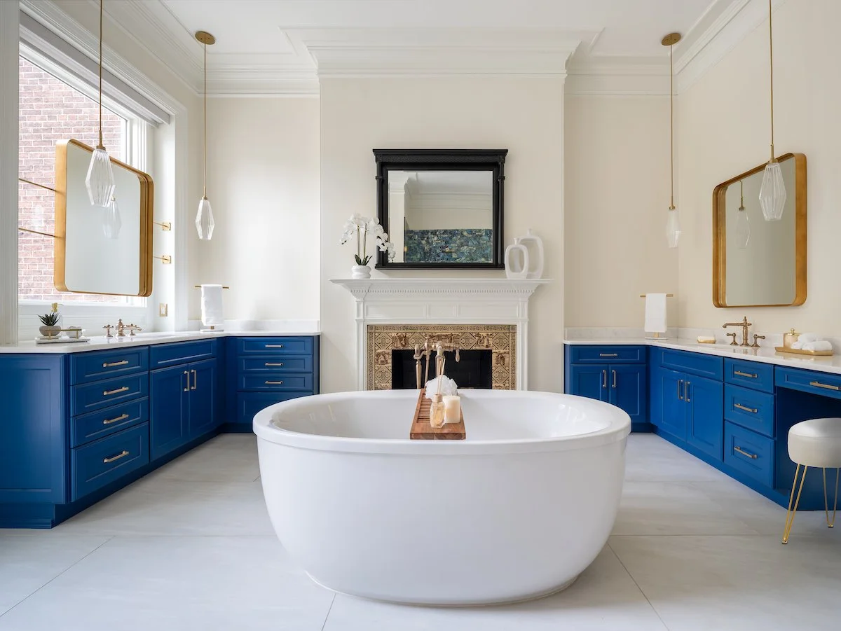 Bright bathroom designed by KD Designs with blue cabinets, a white freestanding bathtub, a fireplace with a mirror above, hanging pendant lights, and large windows.