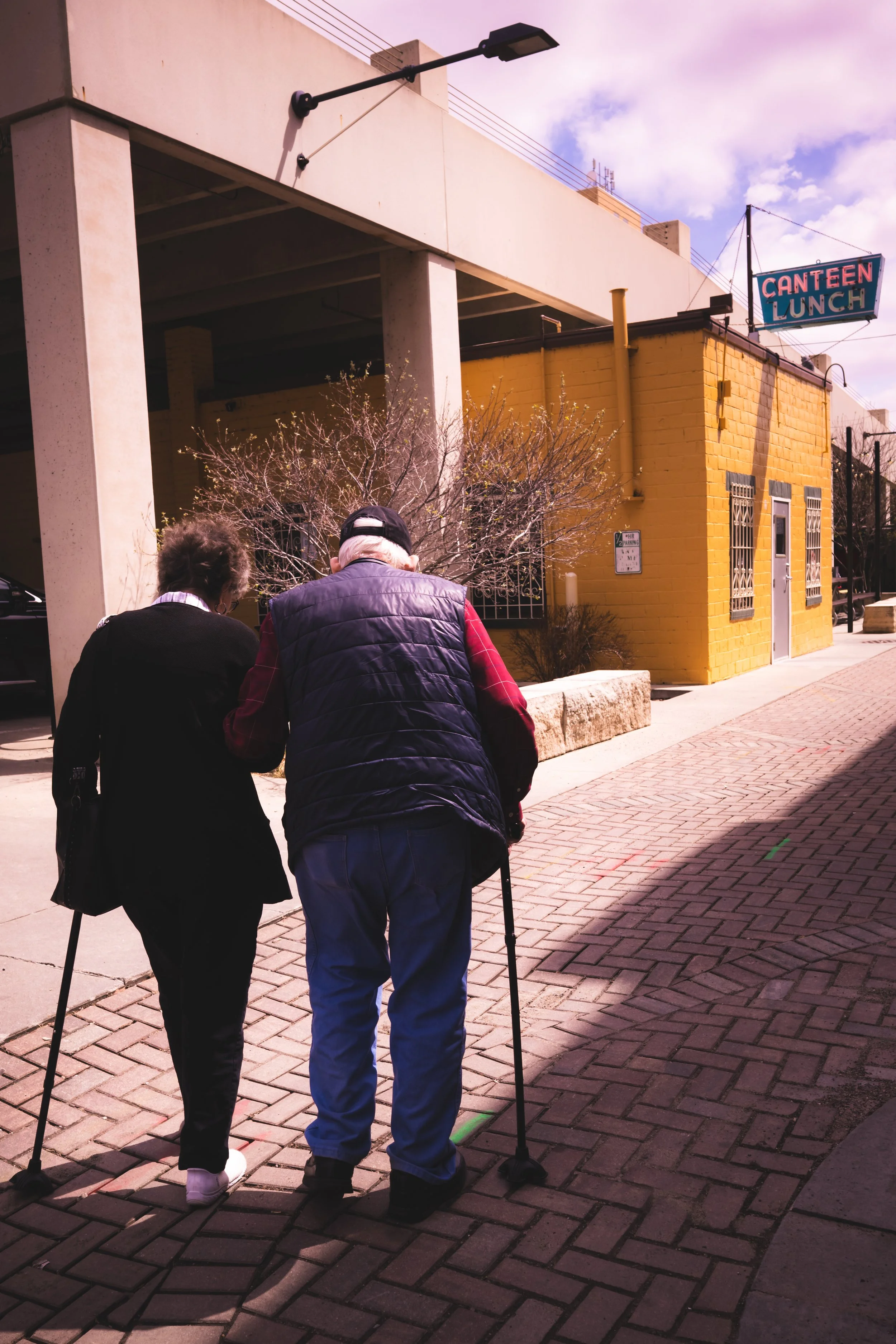Two elderly people, a man and a woman, are walking together with the assistance of canes on a brick sidewalk in front of a yellow brick building with a sign that reads 'CANTEEN LUNCH'. The man is using a cane with a black top and walking stick, while