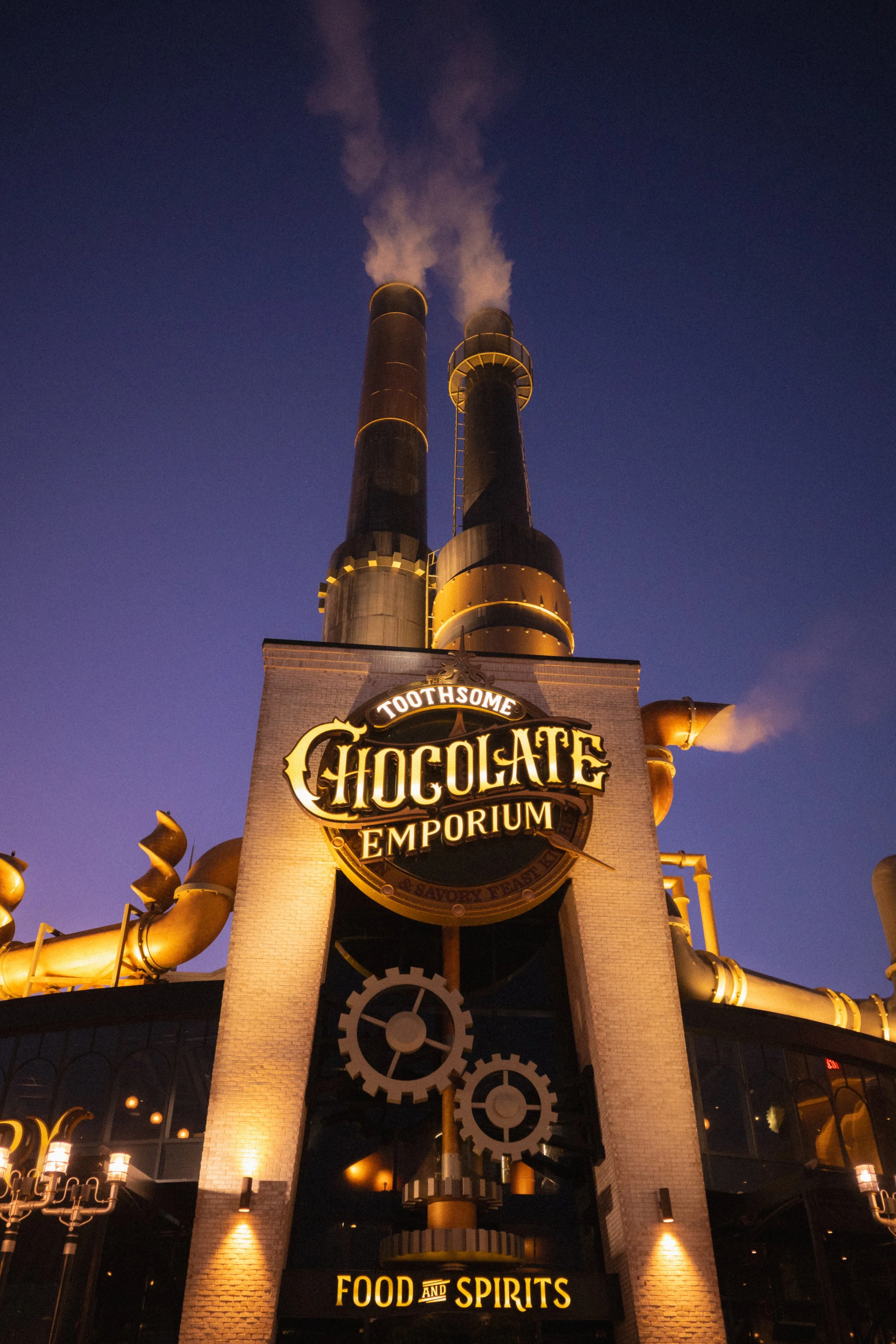 Night view of the Toothsome Chocolate Emporium, a factory-style building with tall black and gold smokestacks emitting smoke, illuminated signage, and gears on the facade.