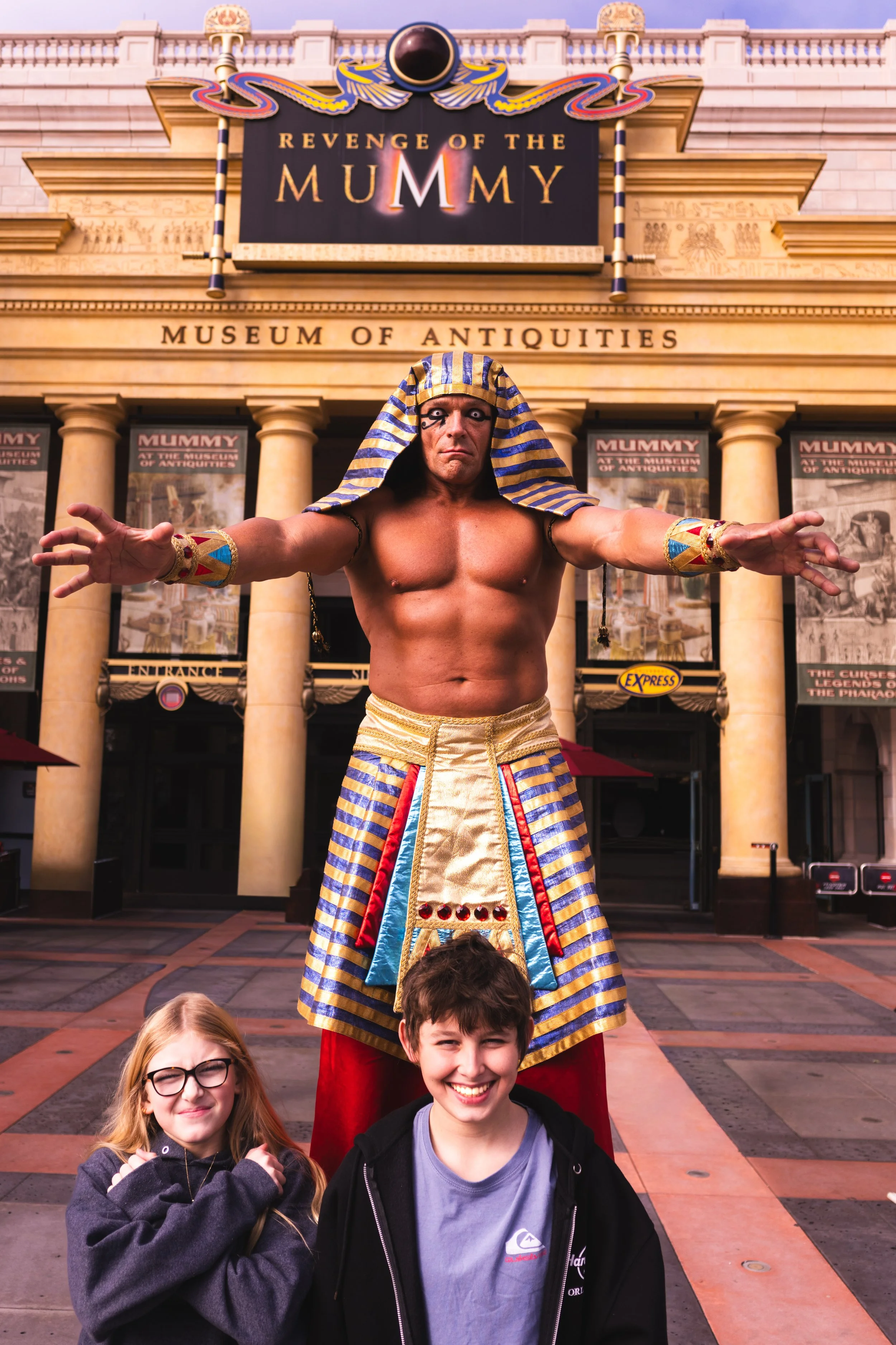 A performer dressed as an ancient Egyptian Pharaoh standing with arms outstretched in front of a Museum of Antiquities building with a promotional sign for 'Revenge of the Mummy.' Two children, a girl with glasses and blonde hair and a boy with dark 