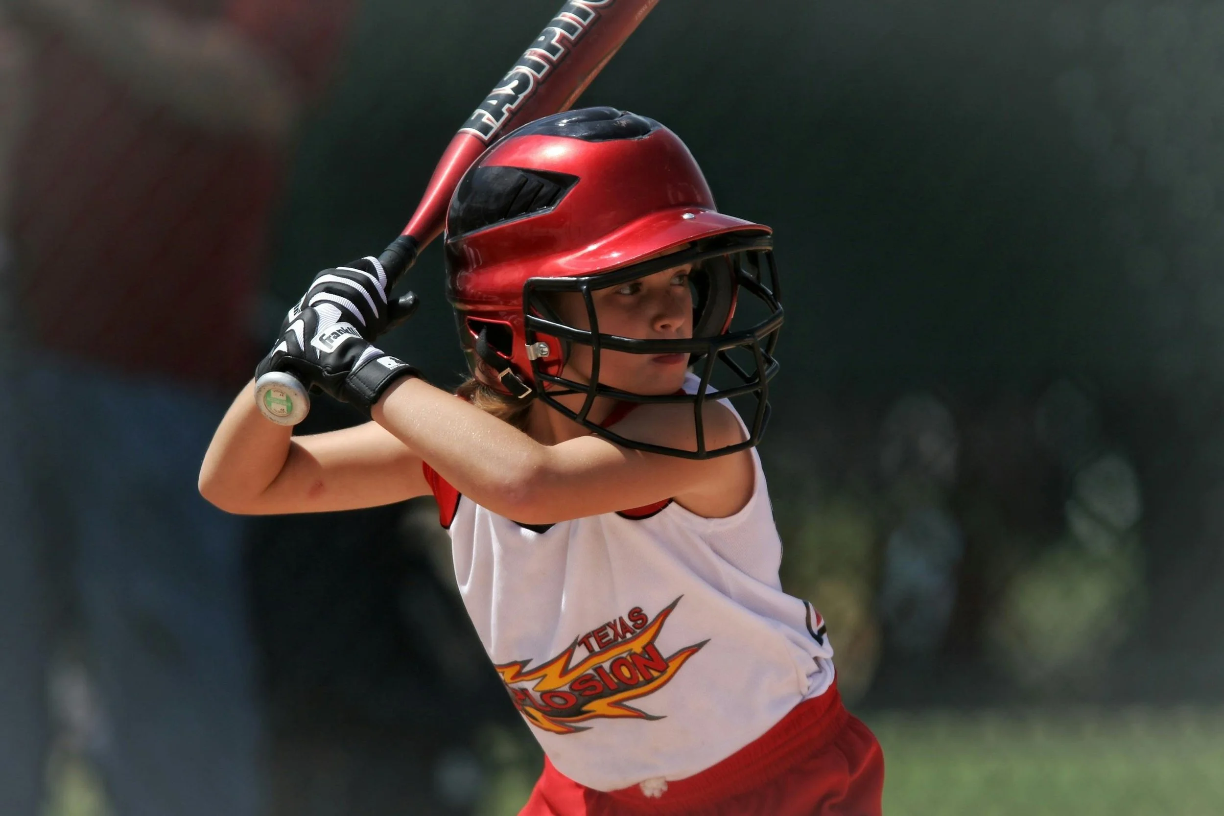 Young girl in a baseball helmet and jersey with Texas Bombers logo, holding a bat ready to swing during a game.