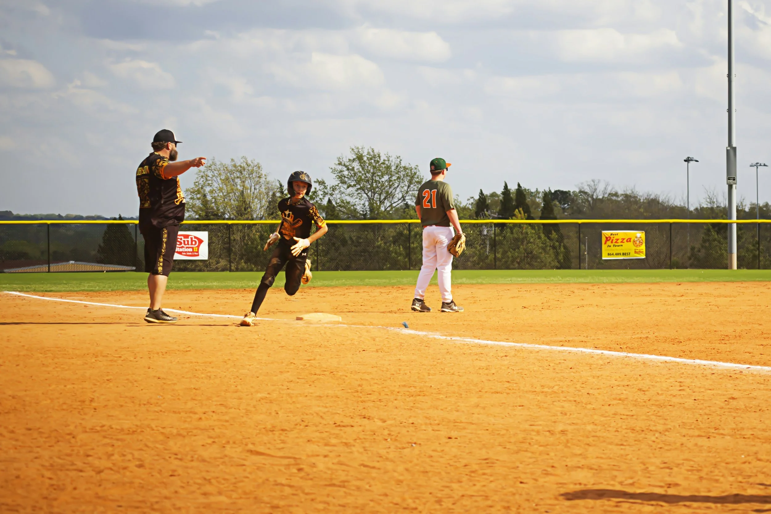 A youth baseball game showing two players, one running towards first base and the other waiting, with a coach pointing and speaking on the field under a partly cloudy sky.