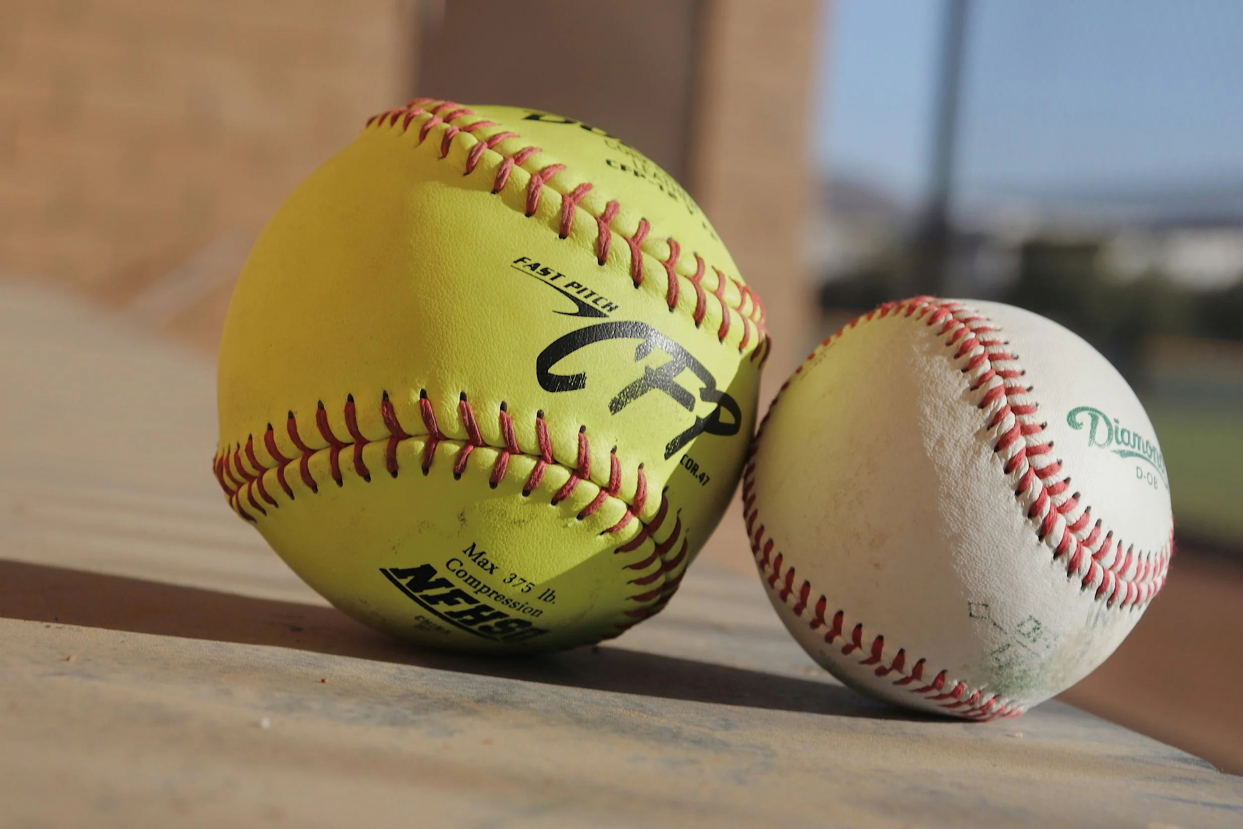 Two baseballs, one yellow with red stitching and black markings, and one white with red stitching and green markings, resting on a wooden surface outdoors.
